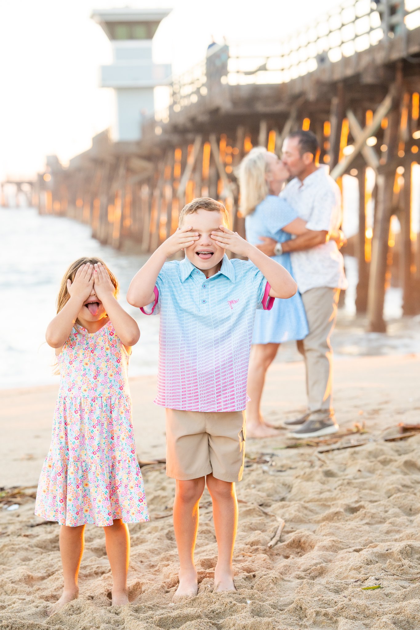 Two children standing barefoot on sandy beach near a wooden pier at sunset, playfully covering their eyes, with a couple embracing and kissing in the blurred background.