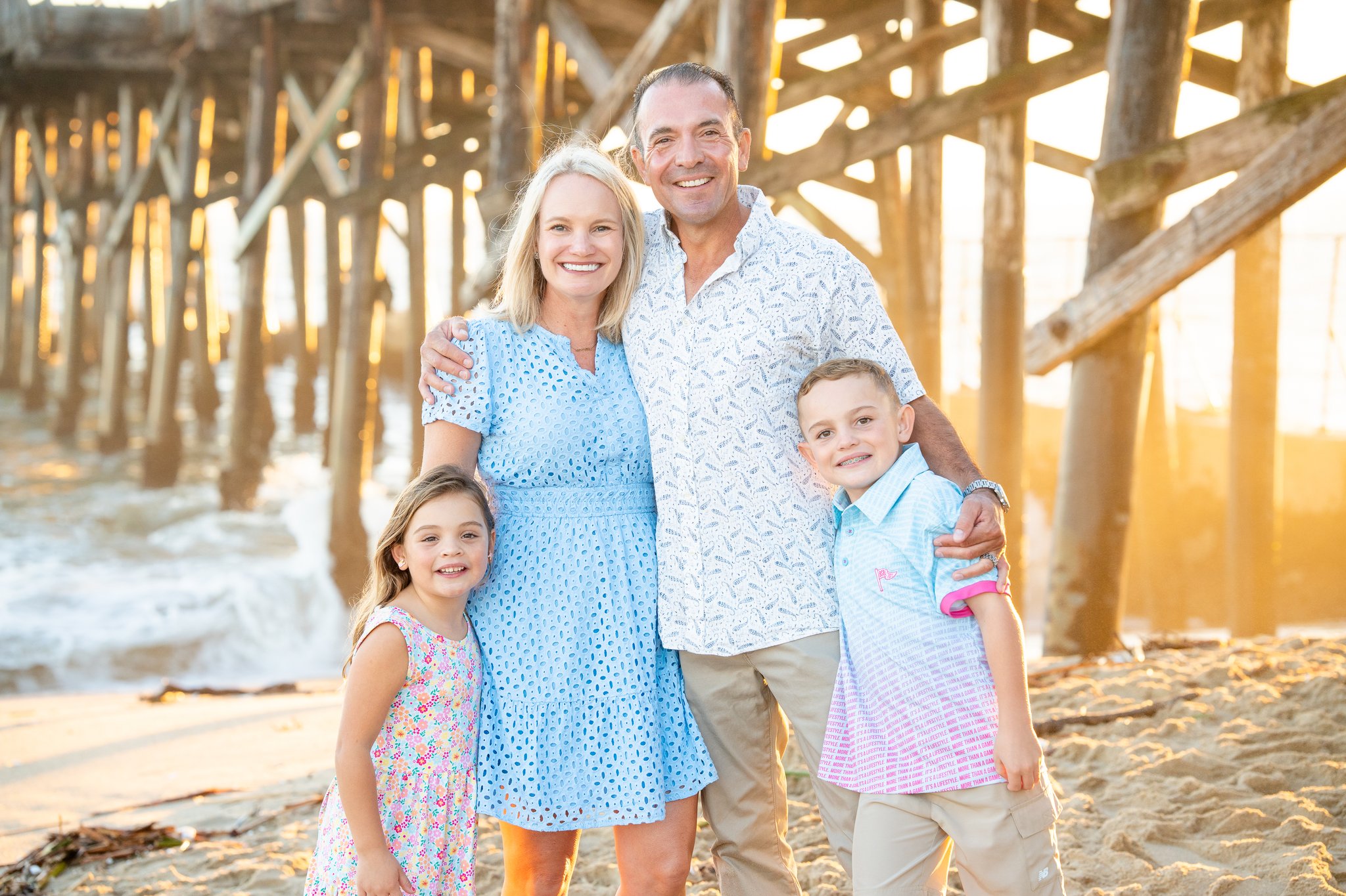 Smiling family of four standing on a sandy beach with a wooden pier structure in the background at sunset, with the parents in the center and children on either side.