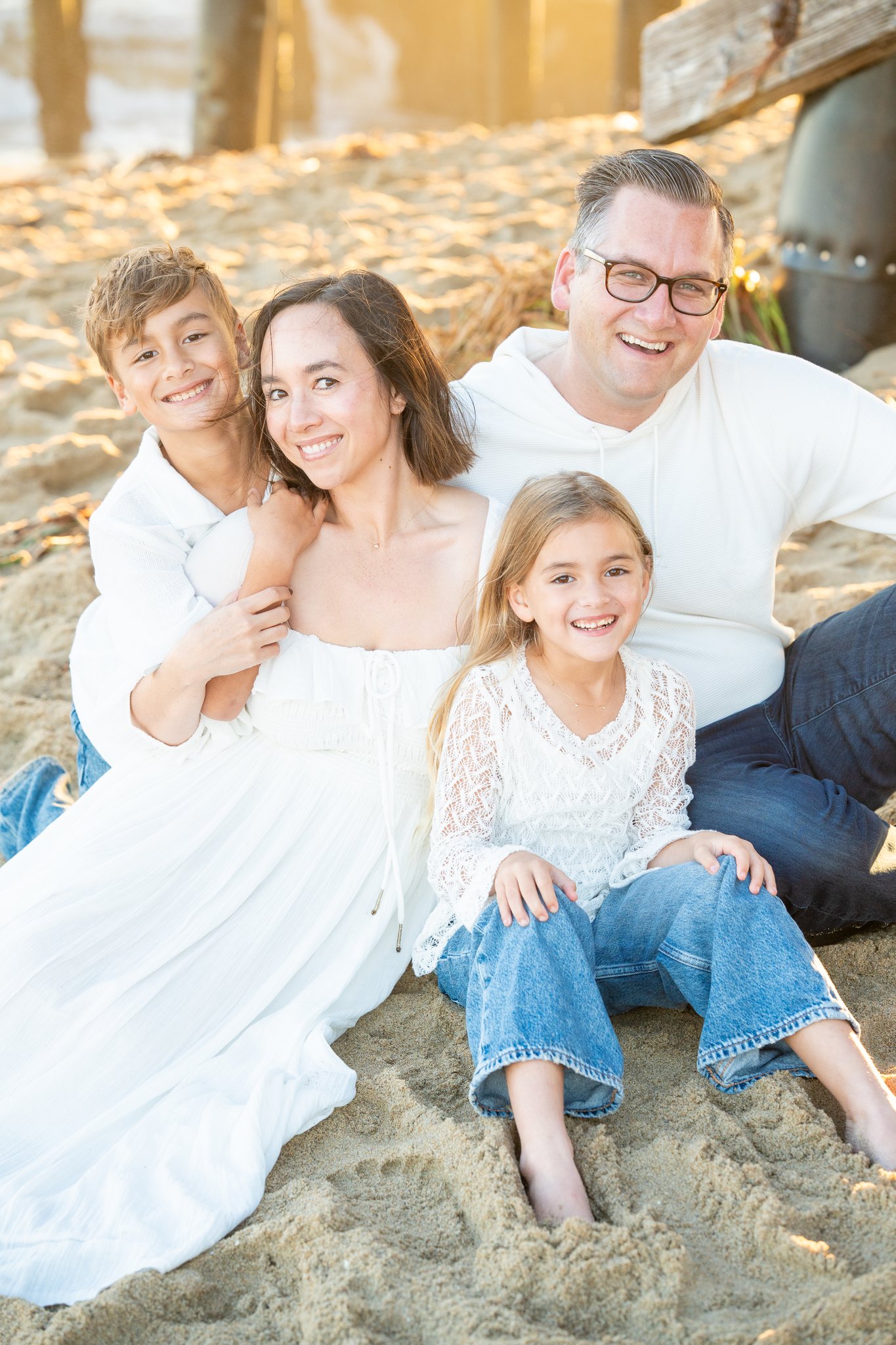 Smiling family of four sitting on sandy beach at sunset, including a mother in a white dress, a father in glasses and white hoodie, a son in white shirt, and a daughter in a white lace top and blue jeans.