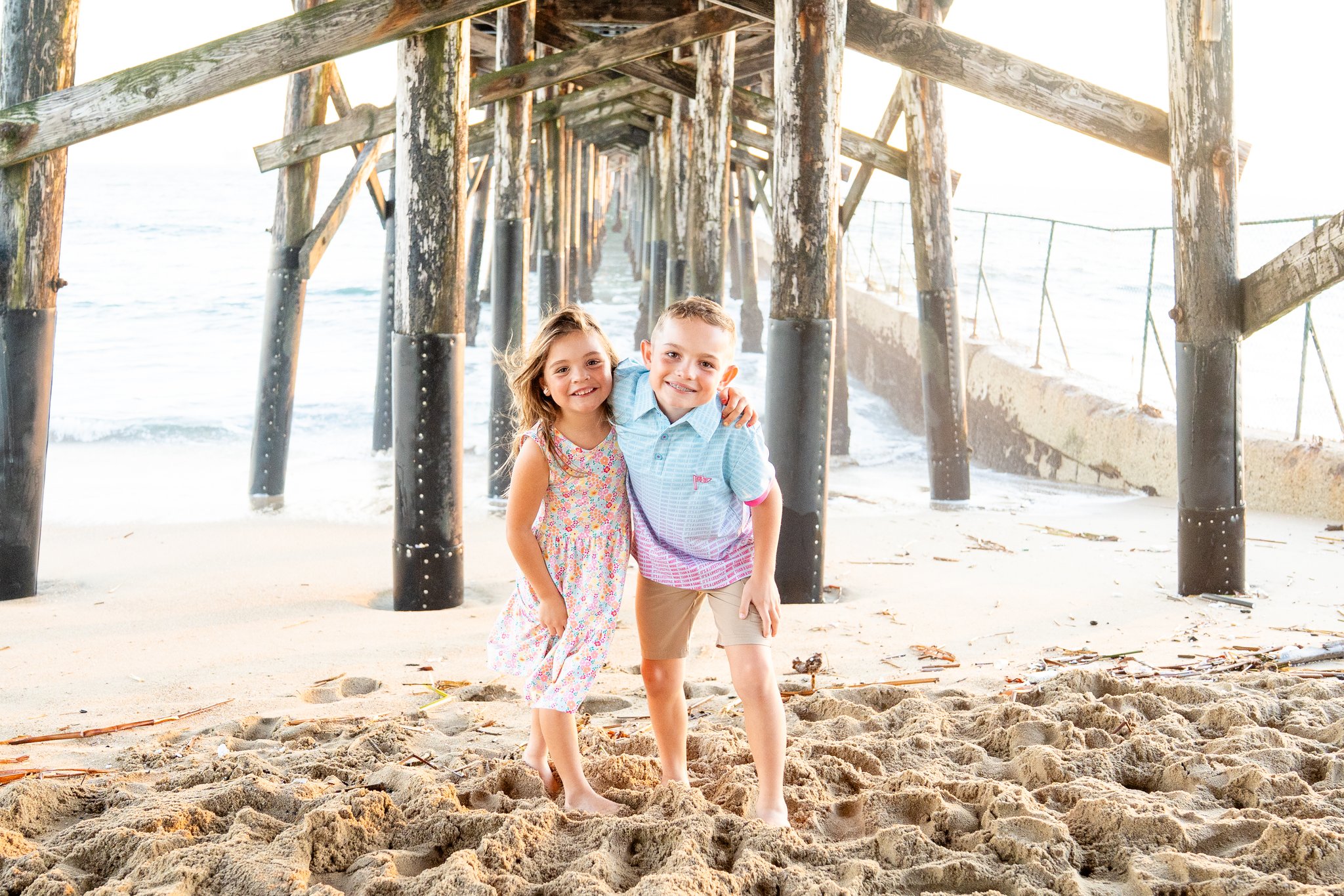 A young girl wearing a floral dress and a young boy in a blue and pink shirt with beige shorts stand barefoot on sandy beach beneath a wooden pier, smiling with the boy's arm around the girl's shoulder.