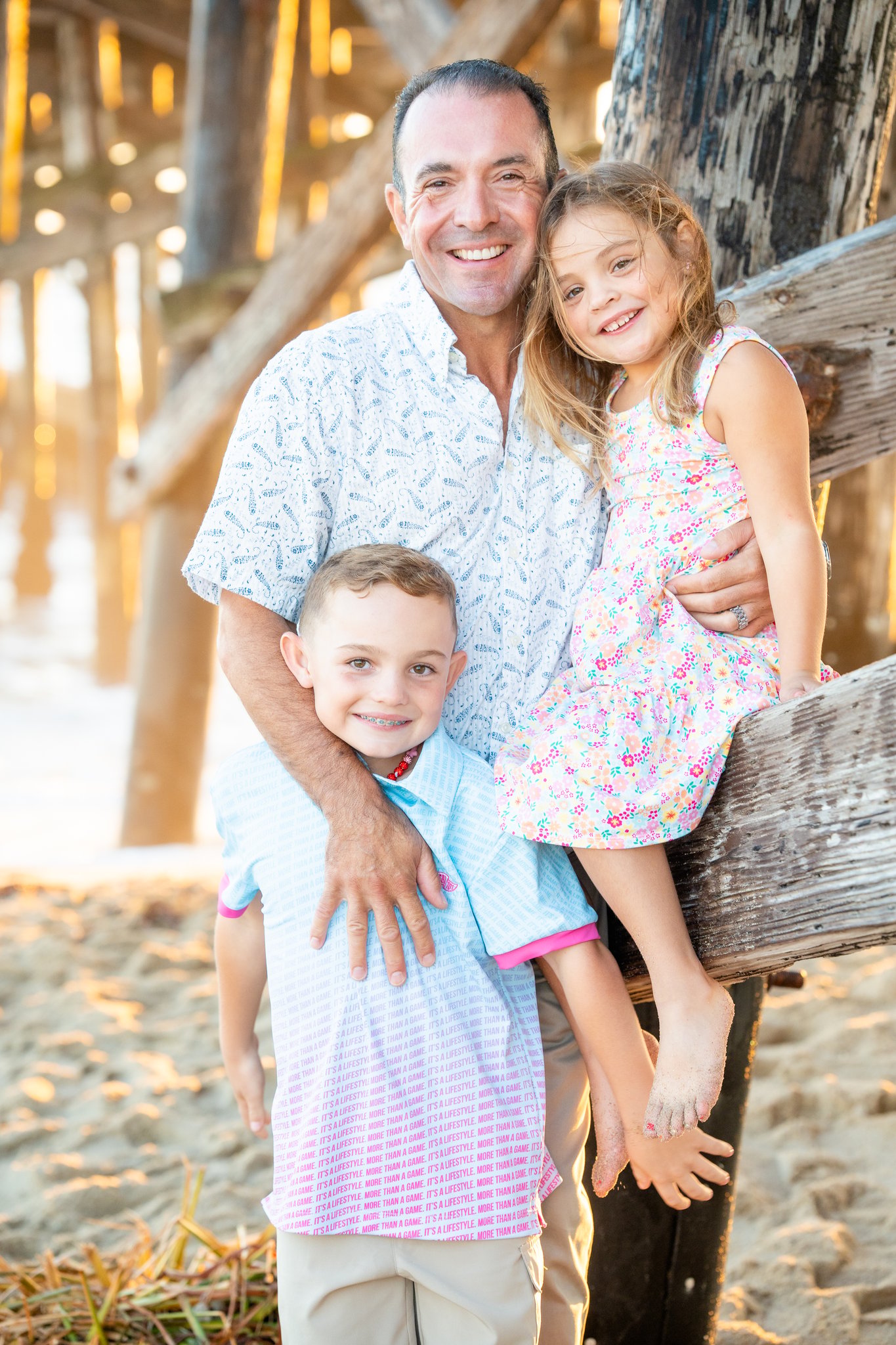 Smiling man in a patterned white shirt hugs two children standing on sandy beach under a wooden pier; the boy with braces and a blue gradient polo shirt stands in front while the girl in a colorful floral dress is held by the man, her bare sand-covered feet visible.