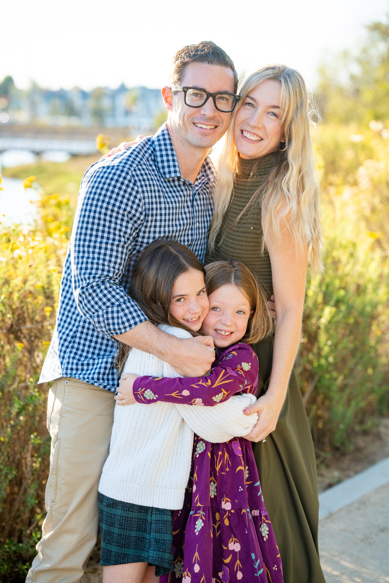 A smiling family of four outdoors with greenery in the background: a man in glasses and a blue checkered shirt, a woman in a green sleeveless dress, and two young girls hugging each other, one wearing a white sweater and green skirt, the other in a purple floral dress.