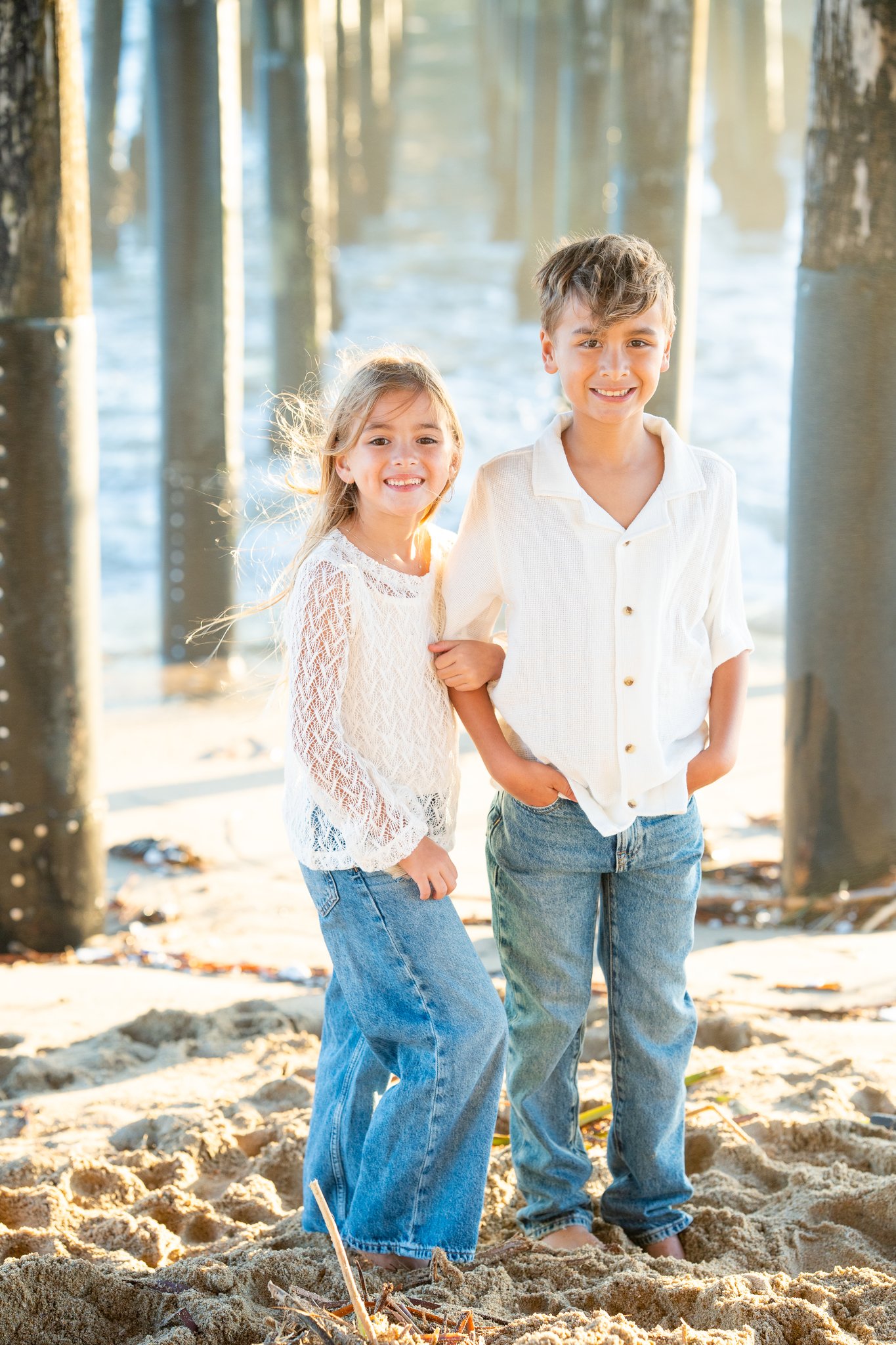 Smiling young boy and girl standing barefoot on sandy beach under wooden pier pillars, both wearing white tops and blue jeans, with ocean water in the background.