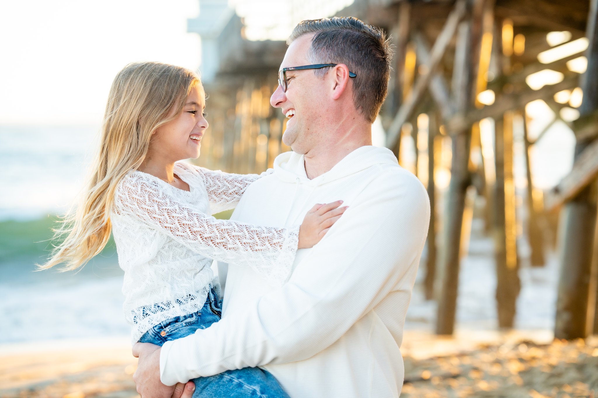 Man wearing glasses and a white hoodie holding and smiling at a young girl in a white lace top and blue jeans on a beach near wooden pier supports, both looking at each other happily.