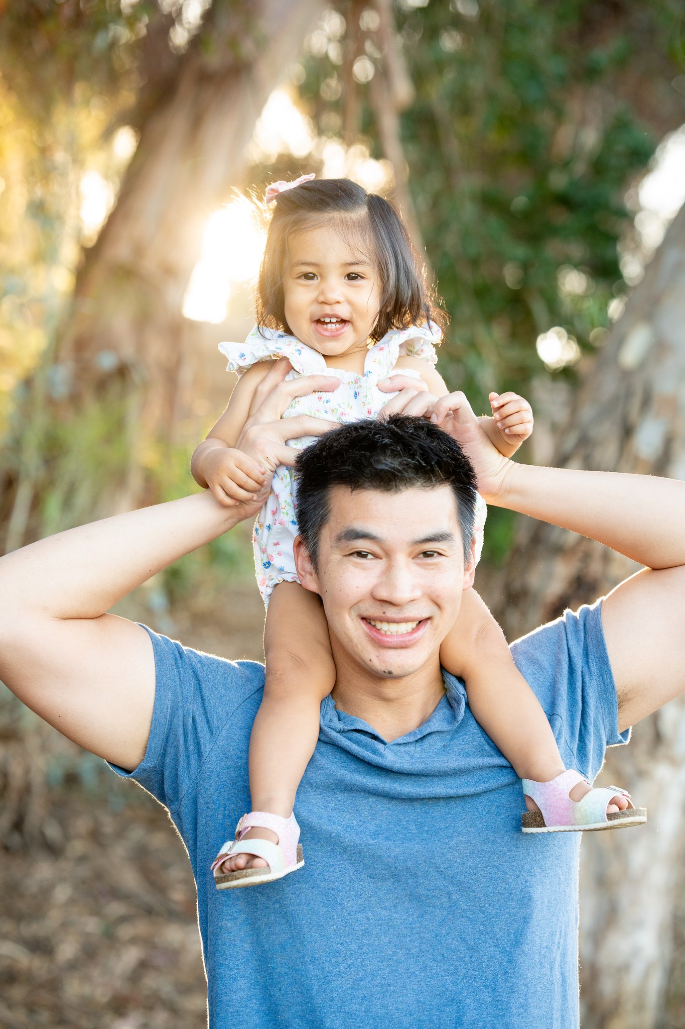 Man wearing a blue shirt smiling with a young girl sitting on his shoulders outdoors, with trees and sunlight in the background.