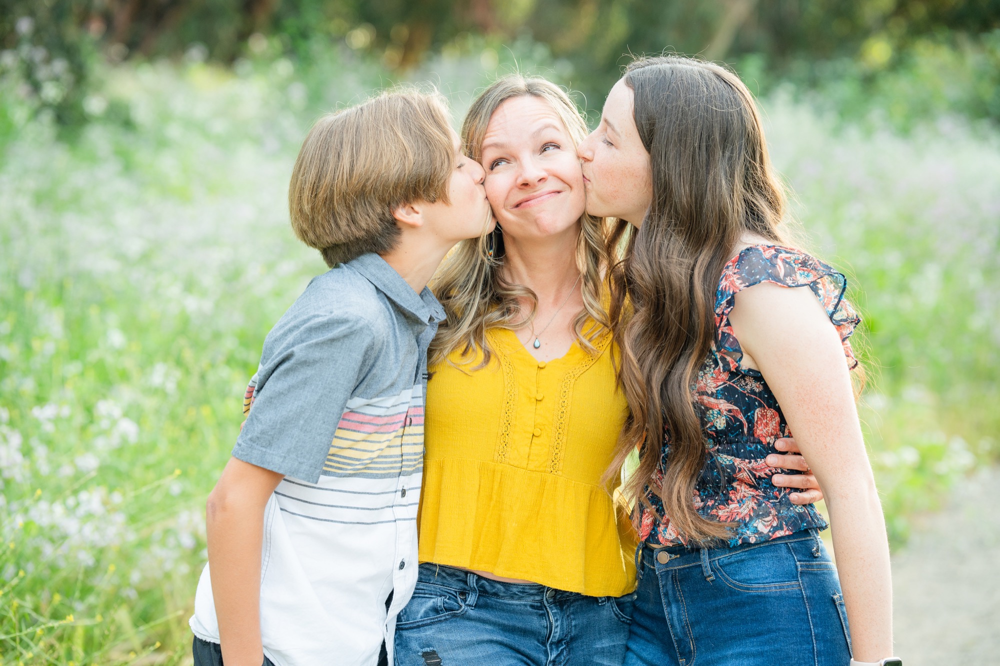 A smiling woman in a yellow blouse is kissed on the cheeks by a boy with short hair in a gray and white shirt on her left and a girl with long brown hair in a floral top and jeans on her right, standing outdoors in a green, floral field.