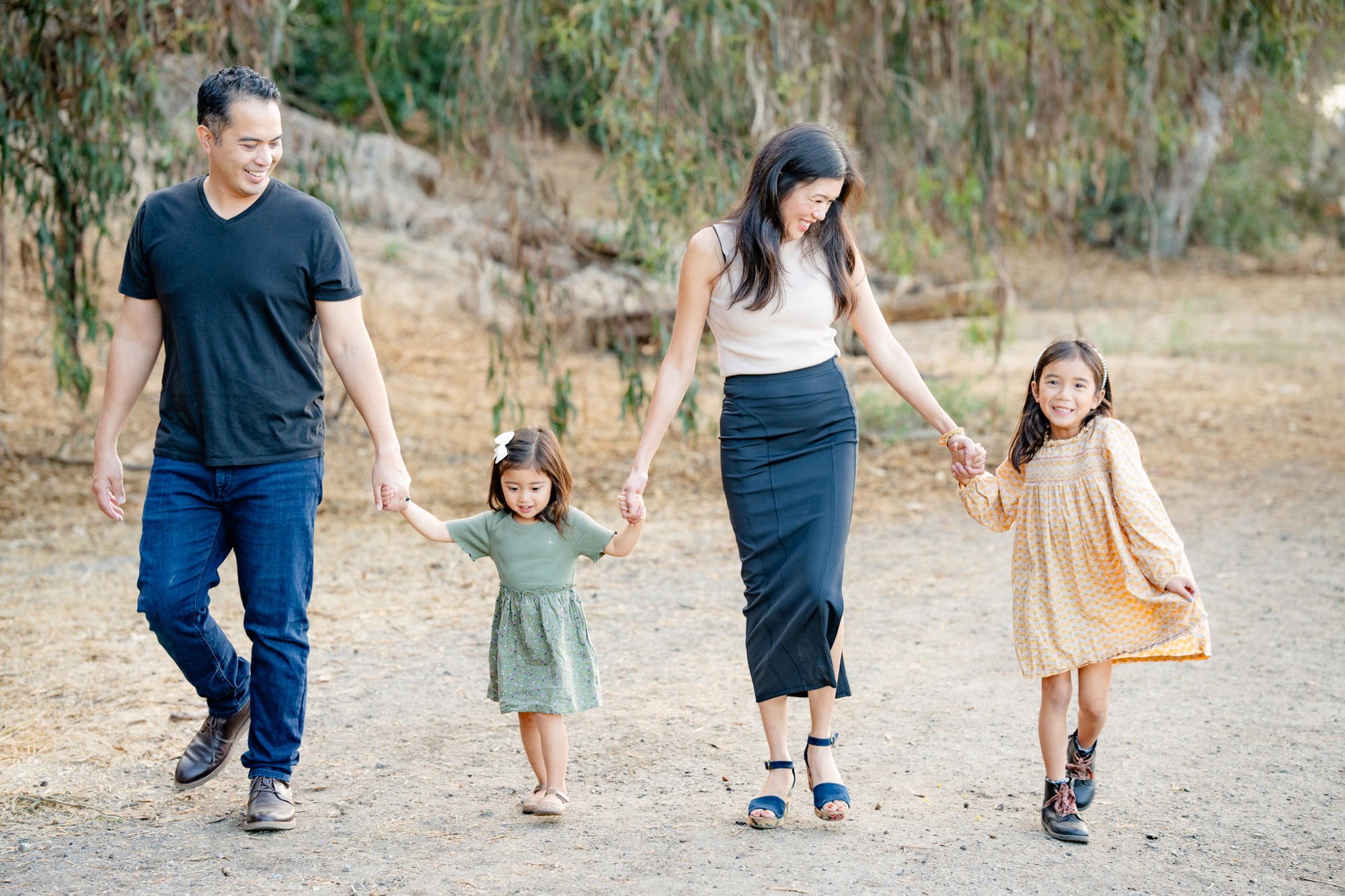 A family of four walking hand in hand outdoors on a dirt path with trees in the background; the father in a black t-shirt and jeans, the mother in a sleeveless white top and black skirt, and their two young daughters wearing dresses and smiling.