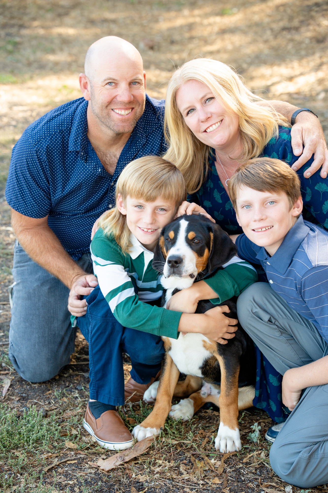A family portrait outdoors with a man and woman standing behind four young boys. The woman wears a green dress with a white vest, and the man wears a beige long-sleeve shirt and blue jeans. The boys wear matching outfits with blue denim shirts, tan pants, and blue shoes. They are smiling and standing on a dirt path with trees and greenery in the background.