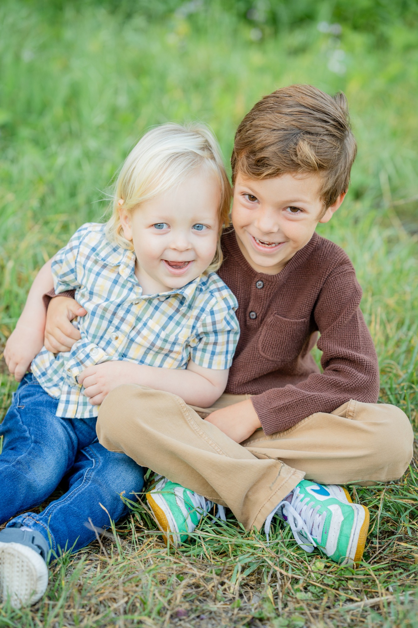 Two young boys sitting on grass, the older boy wearing a brown sweater and khaki pants, hugging the younger boy who has blond hair and wears a plaid shirt and blue jeans, both smiling and looking at the camera.