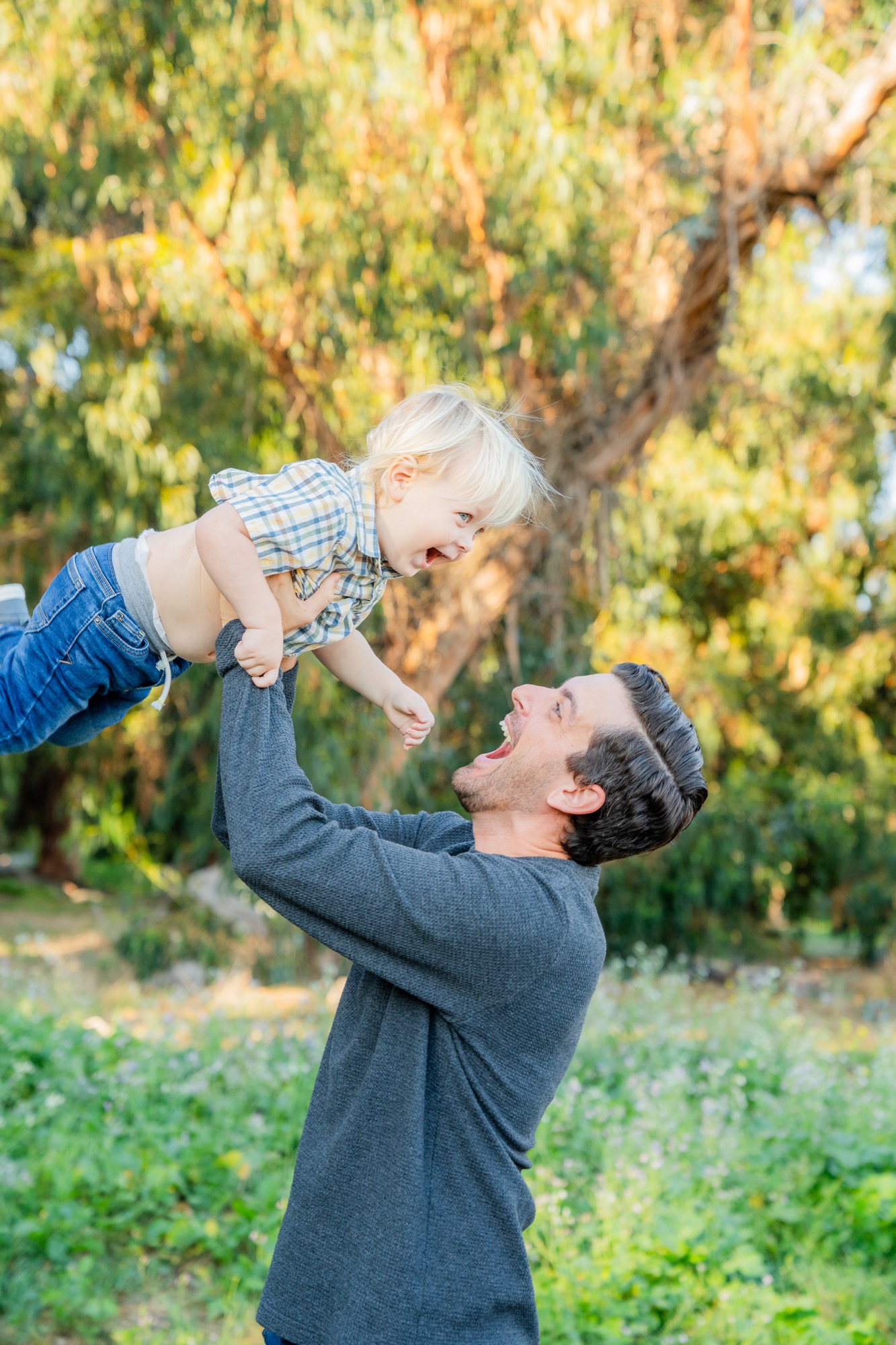 Man with dark hair and beard wearing a gray long-sleeve shirt lifting and playing with a smiling blond toddler in a checkered shirt and jeans outdoors in a green, tree-filled area.