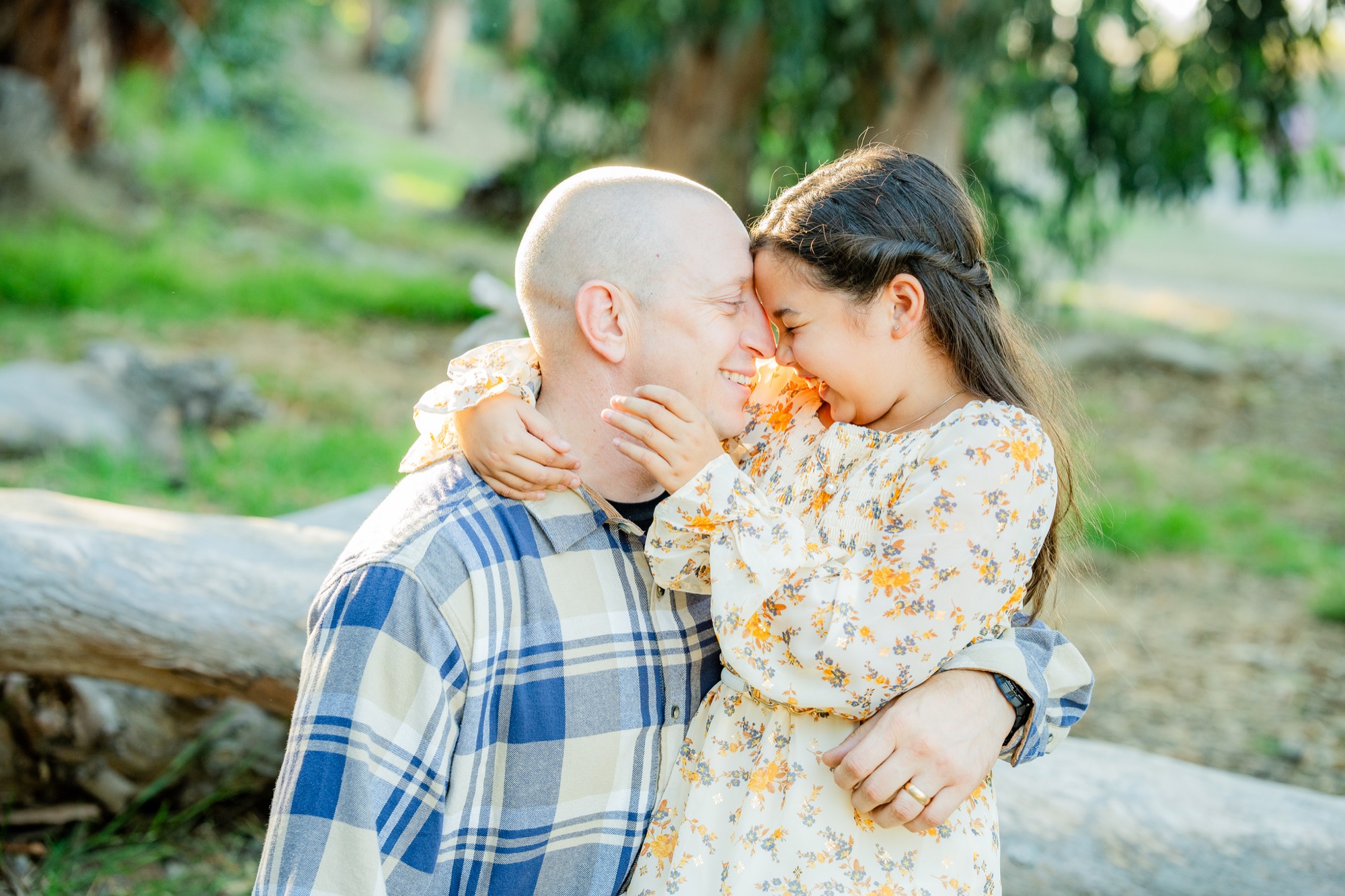 Man in blue and beige plaid shirt embracing and touching foreheads with young girl in a white dress with orange floral pattern, both smiling, outdoors with greenery and a fallen tree in the background.