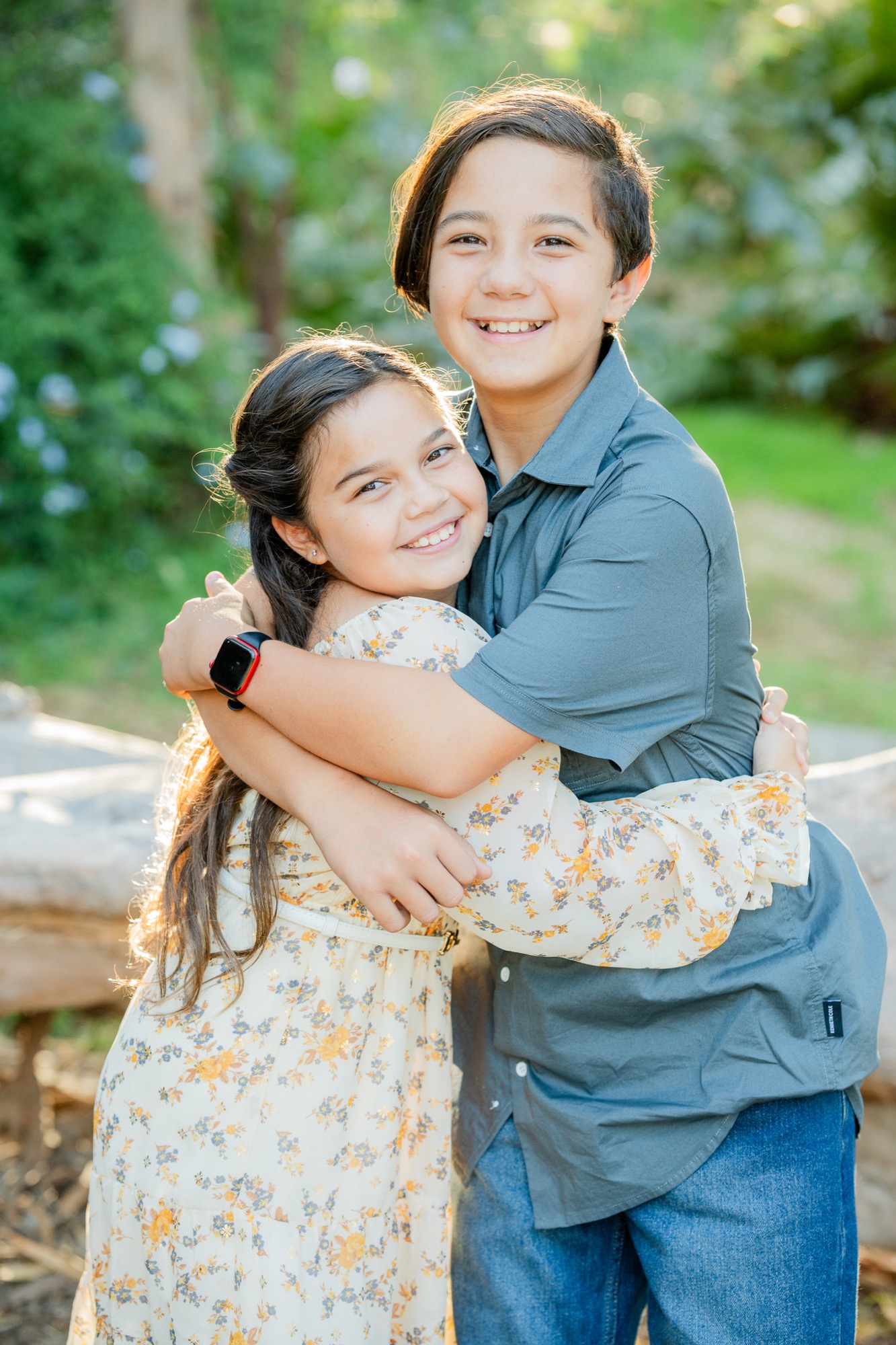 Two smiling children, a boy and a girl, hugging outdoors with green foliage and a large log in the background; the girl wears a light floral dress and the boy wears a blue shirt and jeans.