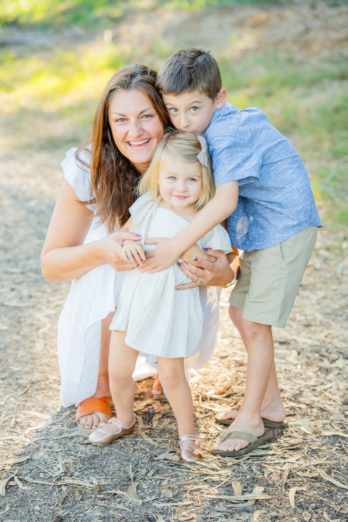 Smiling woman in a white dress crouching outdoors, hugging a young girl in a white dress and a boy in a blue shirt and beige shorts who is hugging the girl from behind.