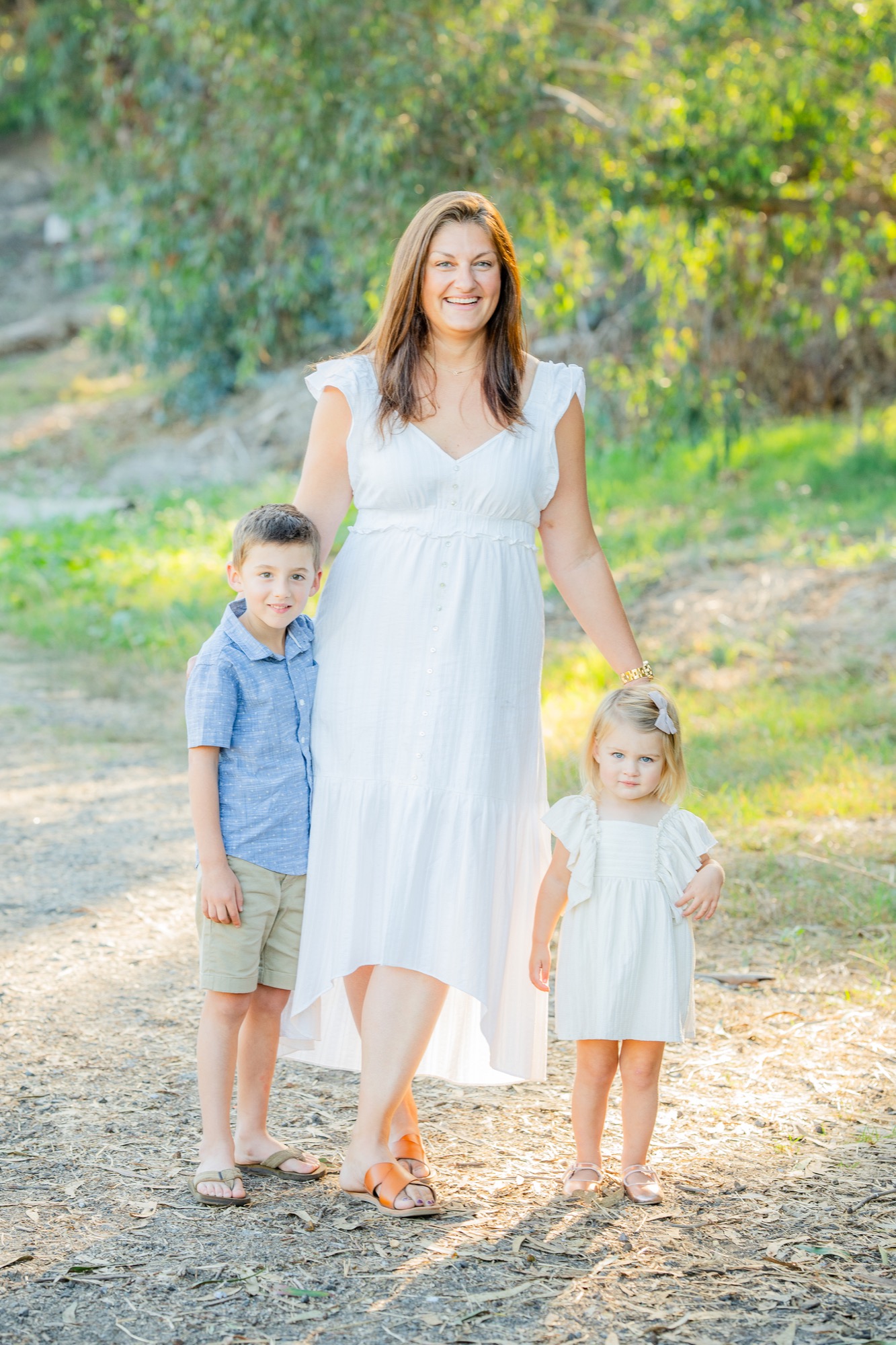 Woman in a white dress standing outdoors on a dirt path with a young boy in a blue shirt and khaki shorts on her left and a young girl in a white dress with a bow in her hair on her right, surrounded by greenery in soft sunlight.