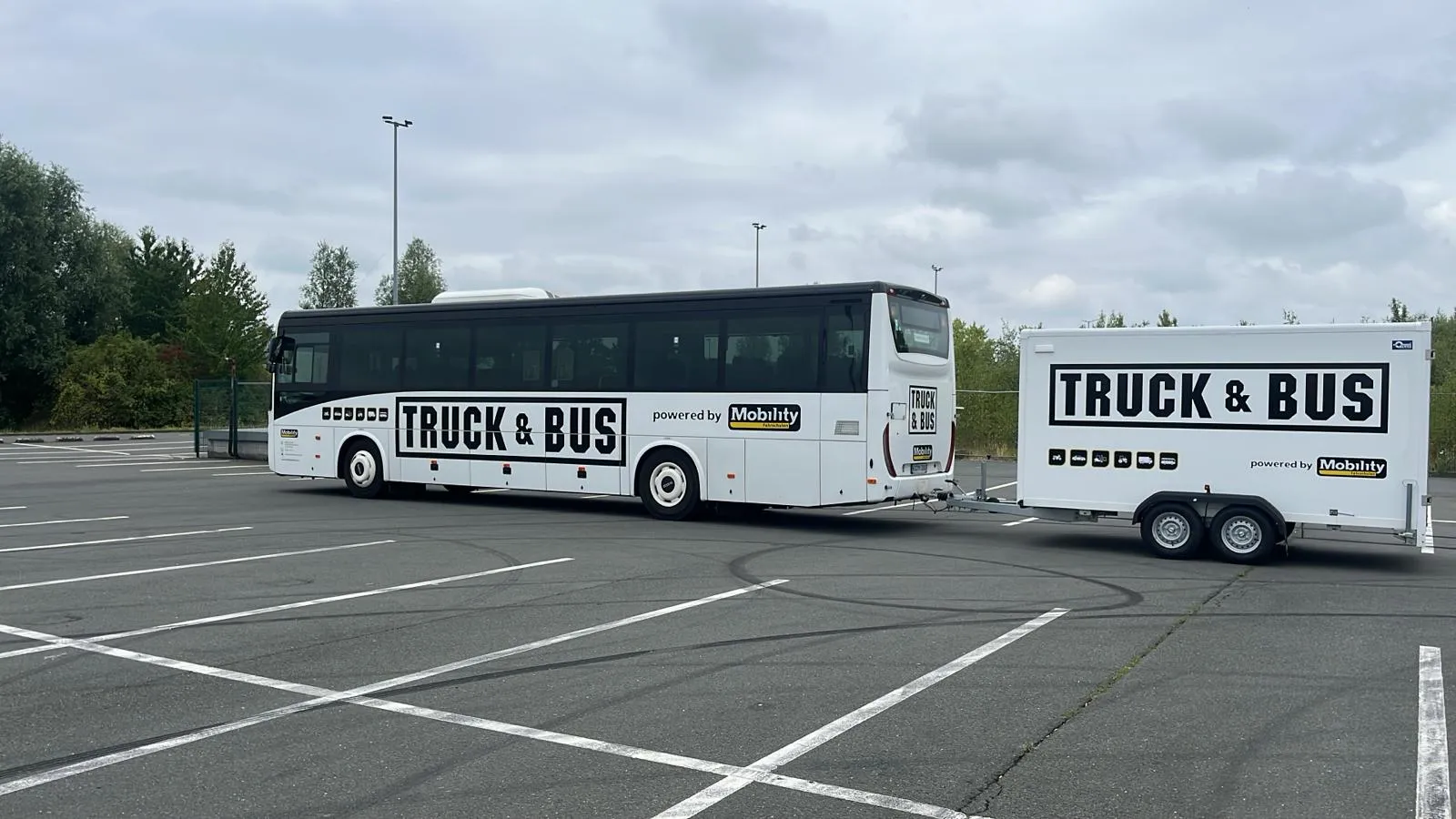 White bus towing a white trailer, both with large black text 'TRUCK & BUS' and 'powered by Mobility' logos, parked in an empty lot under a cloudy sky.