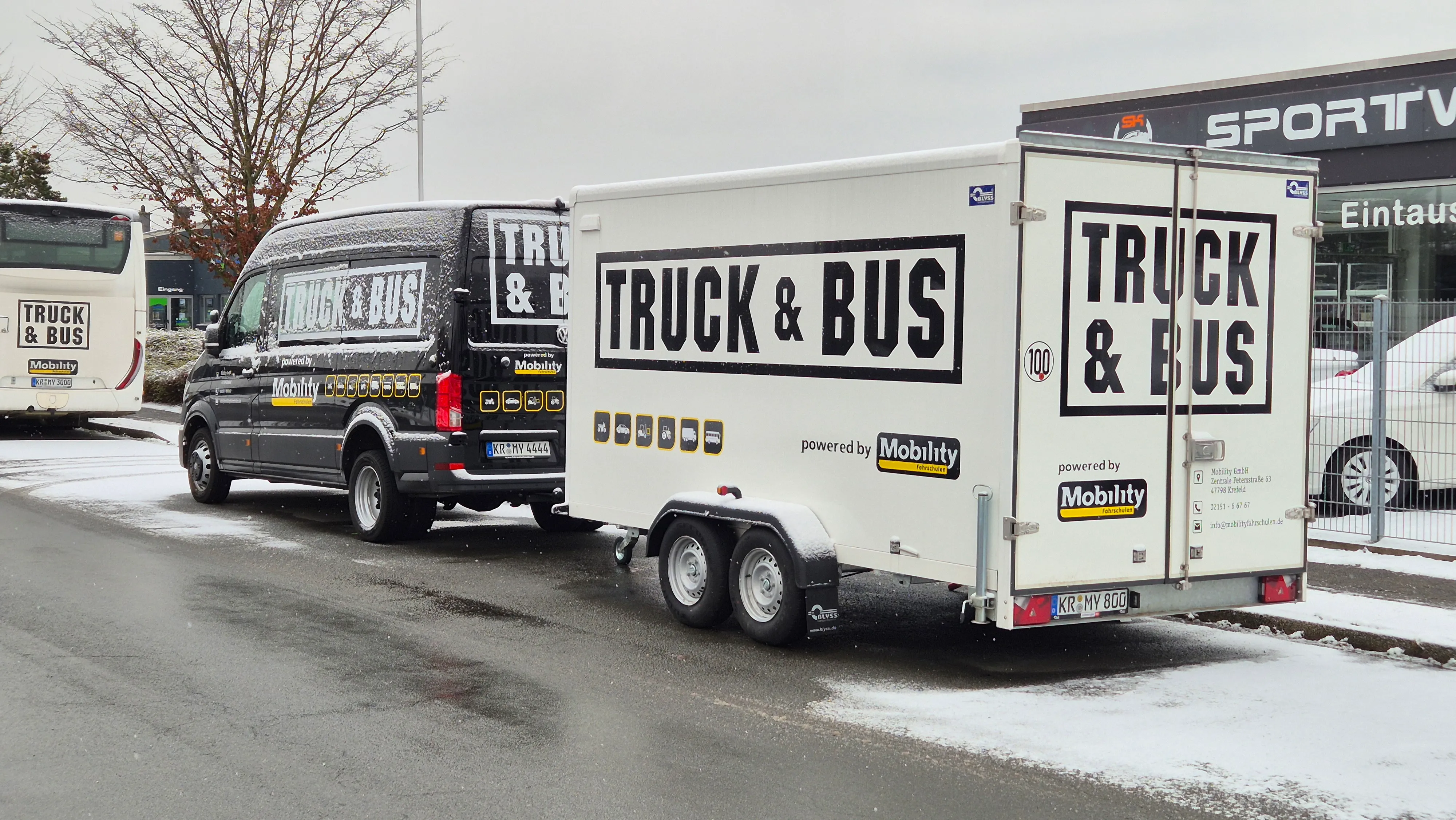 Black van towing a white trailer, both displaying large TRUCK & BUS logos and Mobility branding, parked on a snowy street.