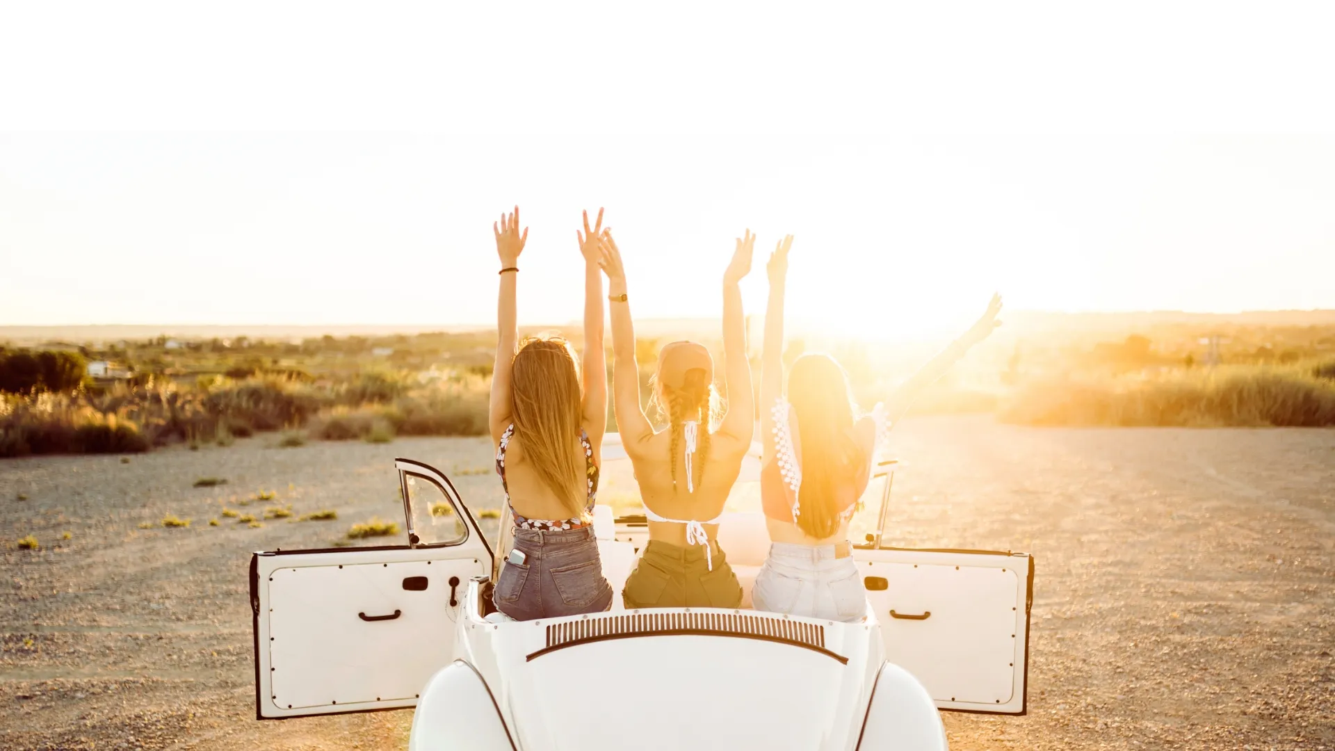 Three young women sitting on the back of a white convertible car with open doors, raising their arms towards a bright sunset.