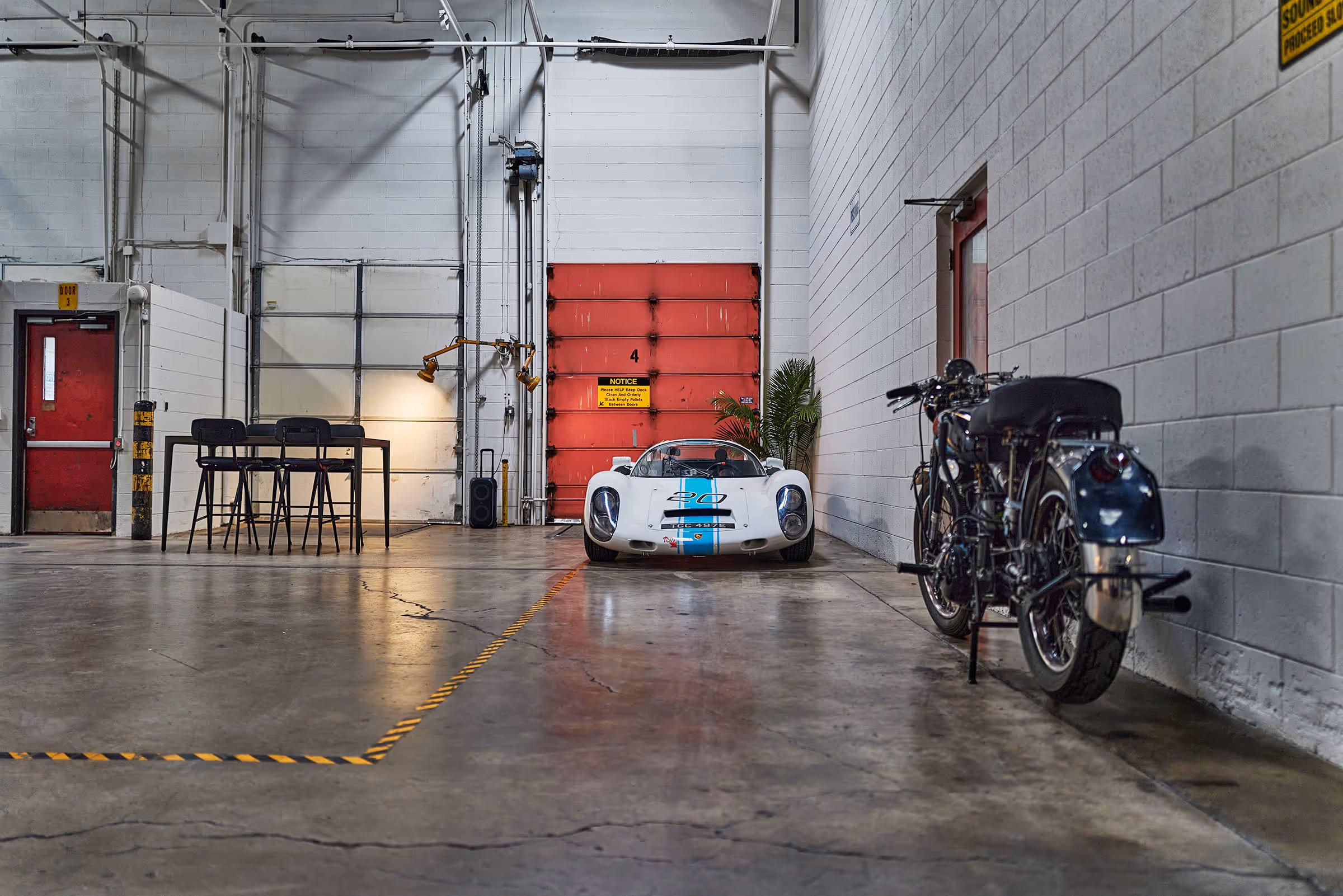 White vintage race car parked inside a spacious industrial garage with a motorcycle nearby and a tall table with chairs on the left.