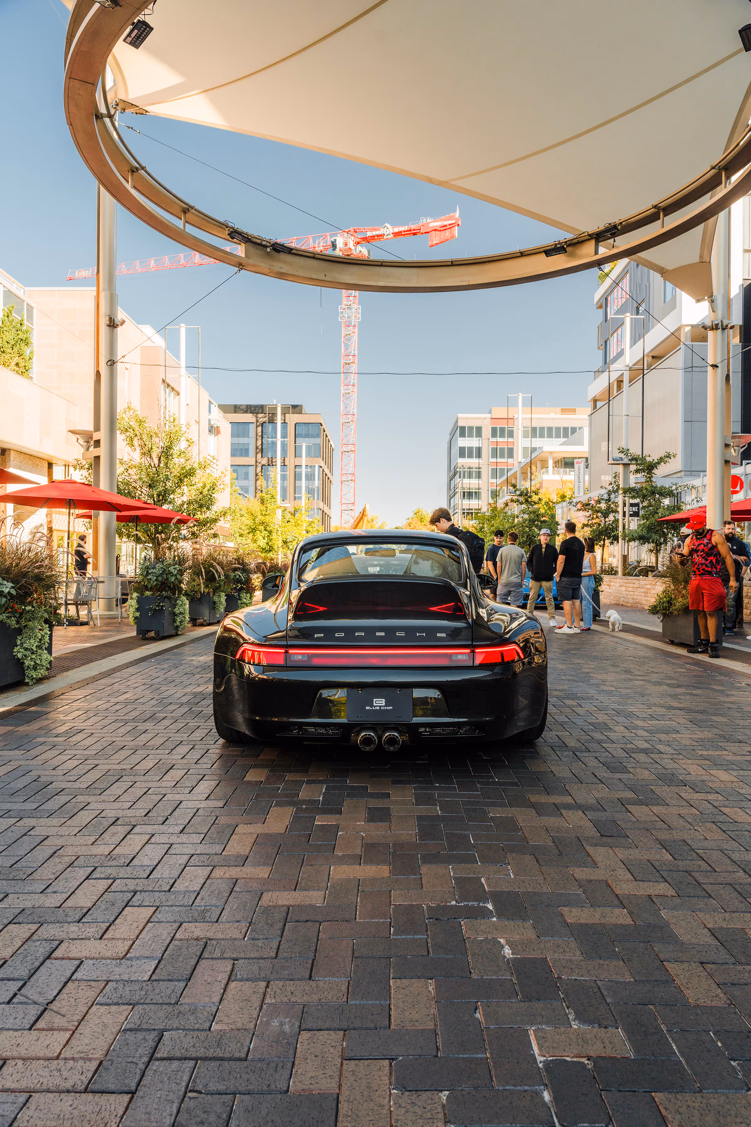 Rear view of a black Porsche parked on a brick-paved street with people standing and walking nearby under a large modern canopy.