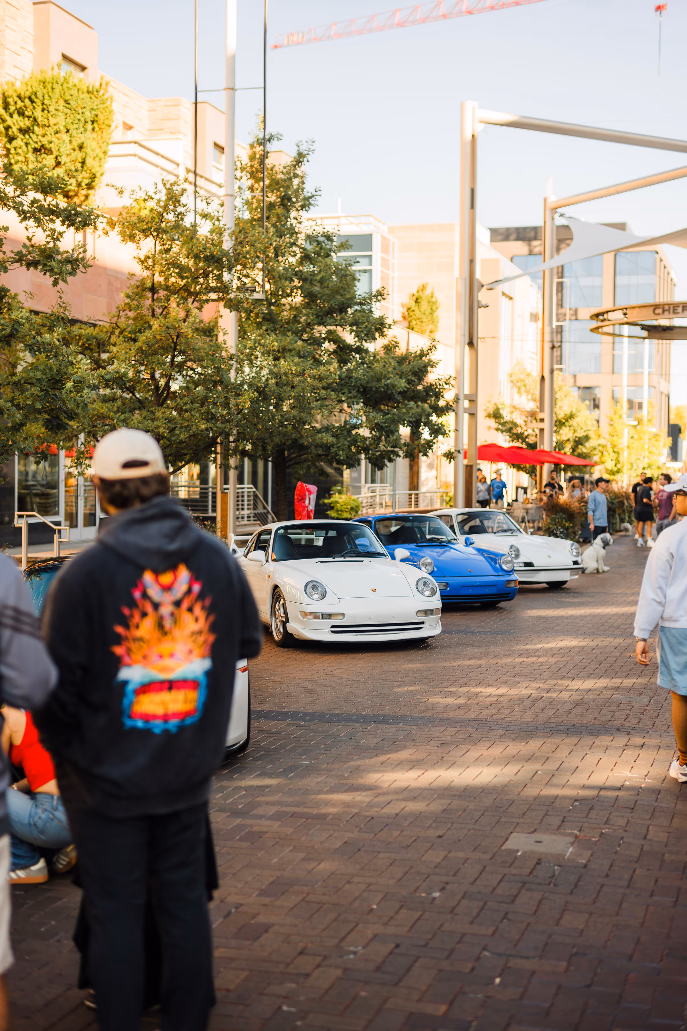 Three vintage Porsche sports cars, two white and one blue, parked on a brick-paved street with trees and buildings in the background.