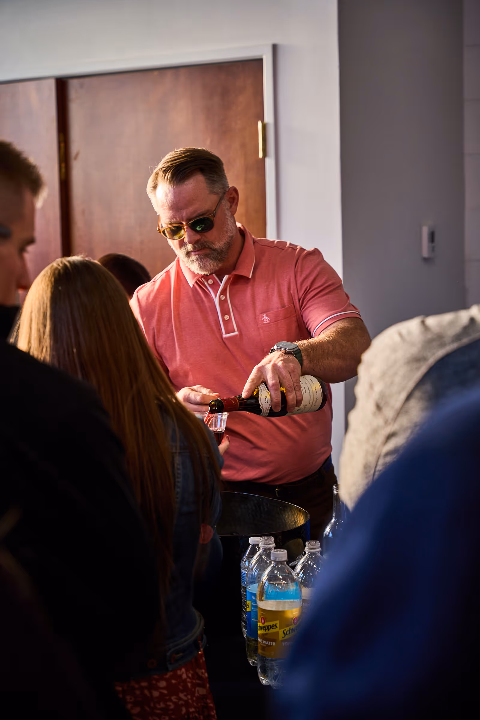Man in pink shirt and sunglasses pouring red wine into a plastic cup held by a woman in a crowded indoor setting.