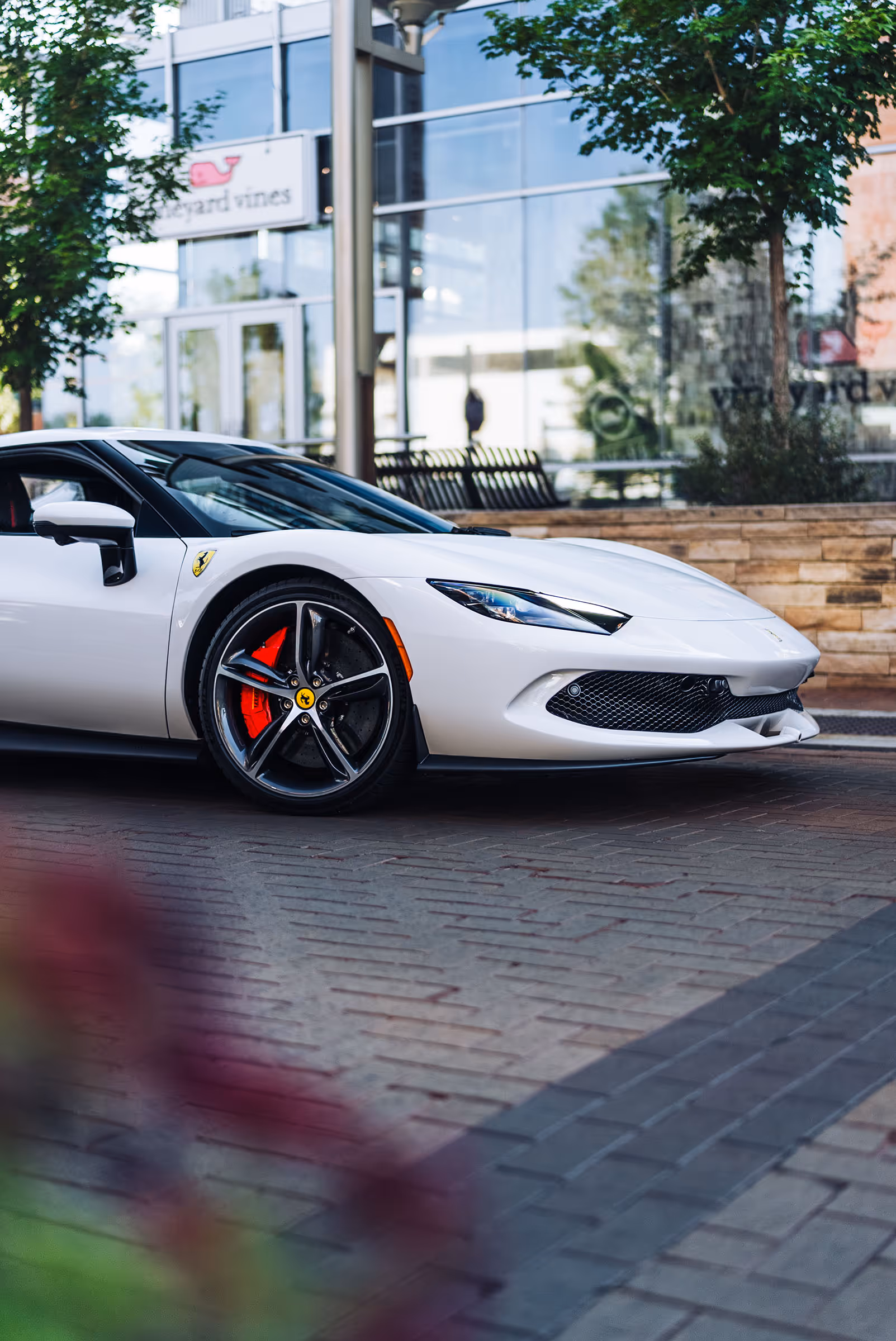 Side view of a white Ferrari sports car parked on a brick street with trees and storefront in the background.