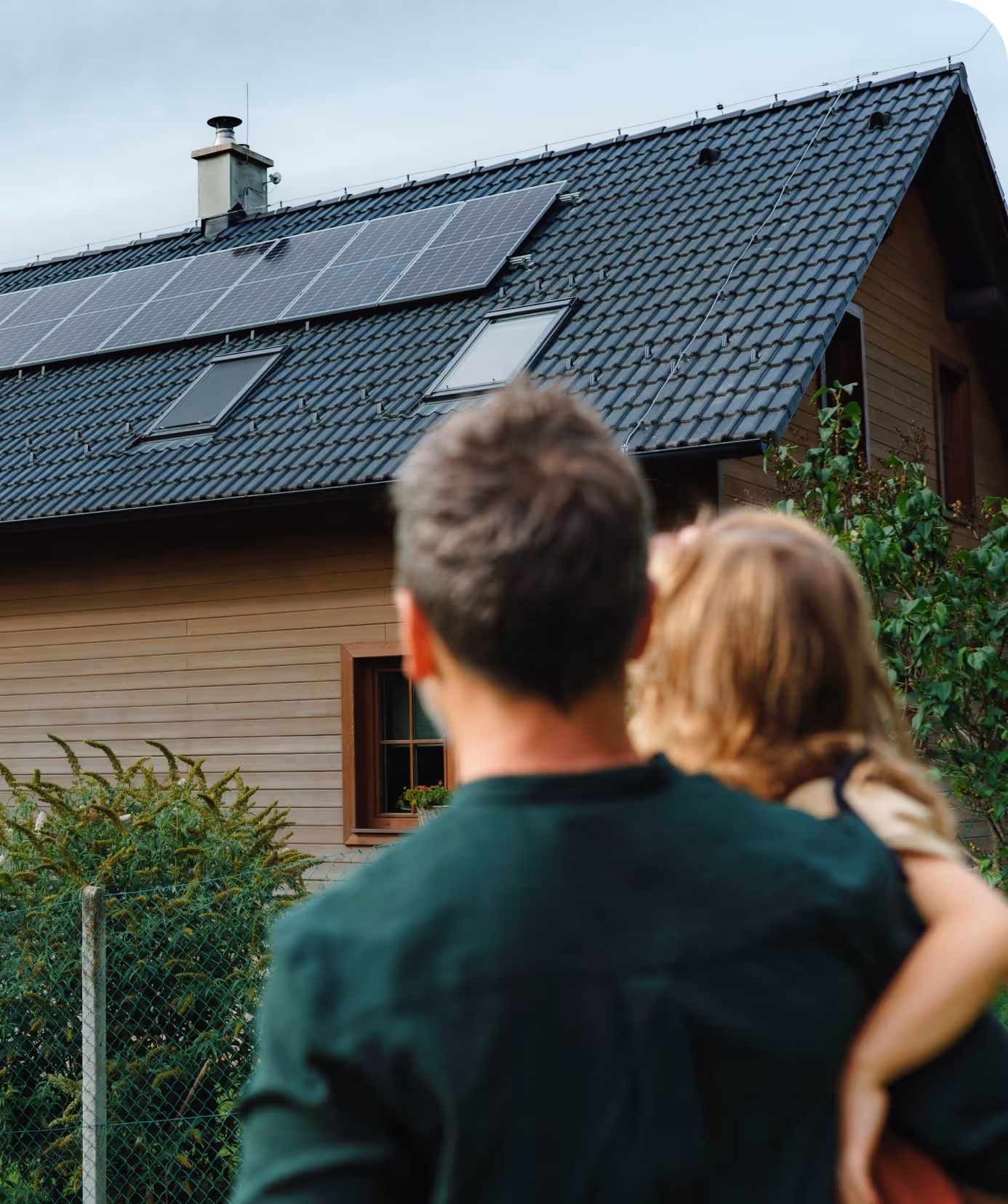 Padre con su hija en brazos de espaldas mirando a las placas solares del tejado de su casa.