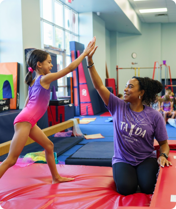 Young girl in a pink leotard giving a high-five to a female gymnastics coach wearing a purple T-shirt indoors on a red mat.