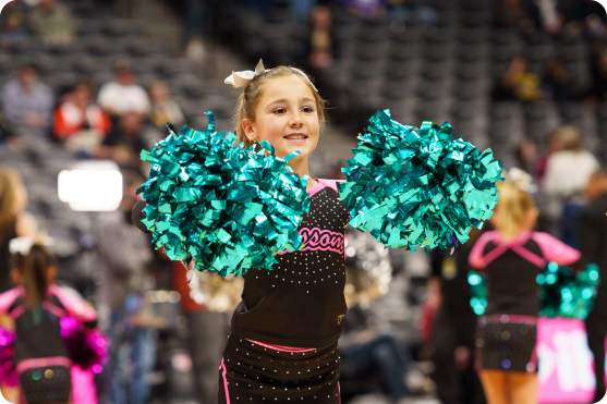 Young cheerleader in black outfit with teal pom-poms smiling during a performance indoors.