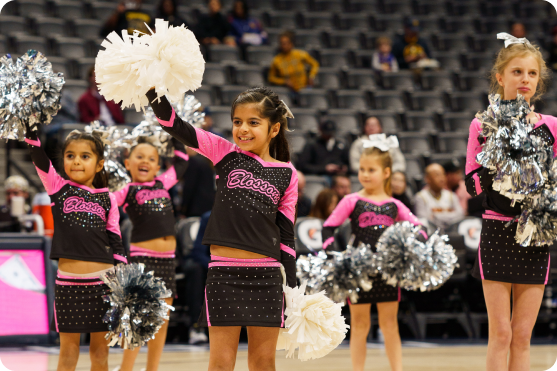 Young girls in black and pink cheerleading outfits performing on a basketball court with silver and white pom-poms.