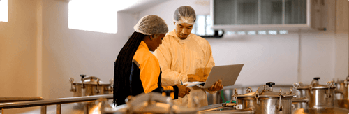 Two workers in hairnets and protective clothing review data on a laptop in an industrial facility.