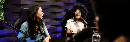 Two women seated at a podcast table with microphones, smiling and engaged in conversation in a studio with a textured wall background and purple lighting.