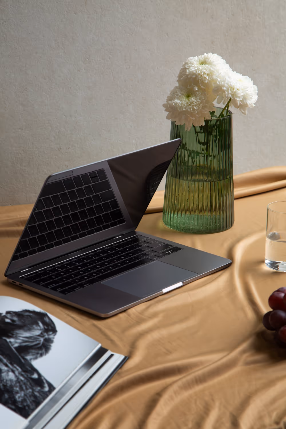 Partially open laptop on a tan cloth next to a green vase with white flowers, a glass of water, grapes, and an open magazine.