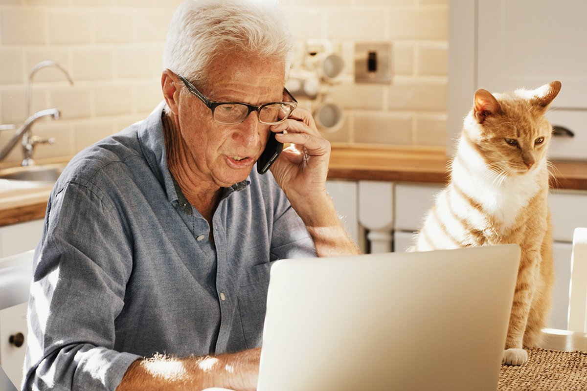 Older man using a laptop and talking on the phone at home with his cat nearby.
