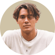 Young man with tousled brown hair wearing a white shirt and a necklace, looking at the camera with a neutral expression.