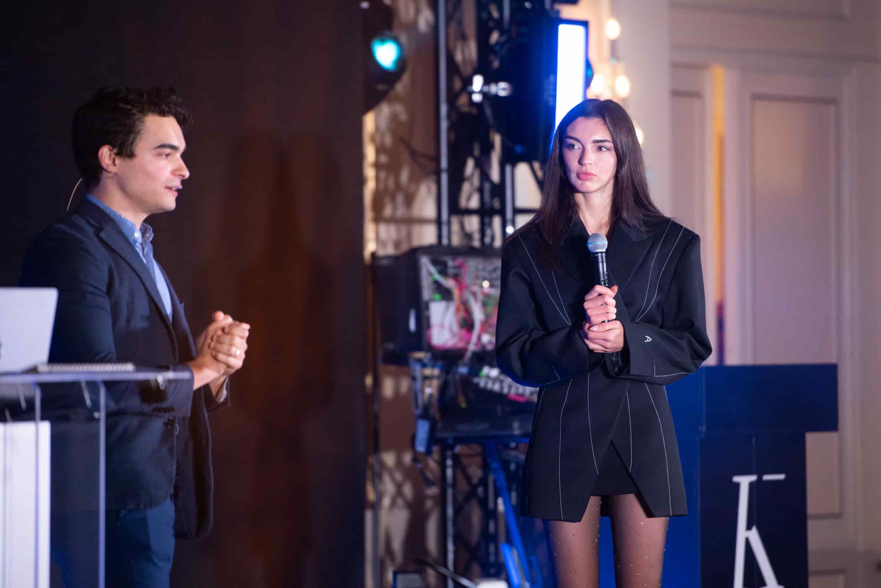 A woman in a black blazer dress holding a microphone stands next to a man in a dark suit speaking on stage with lighting and technical equipment in the background.