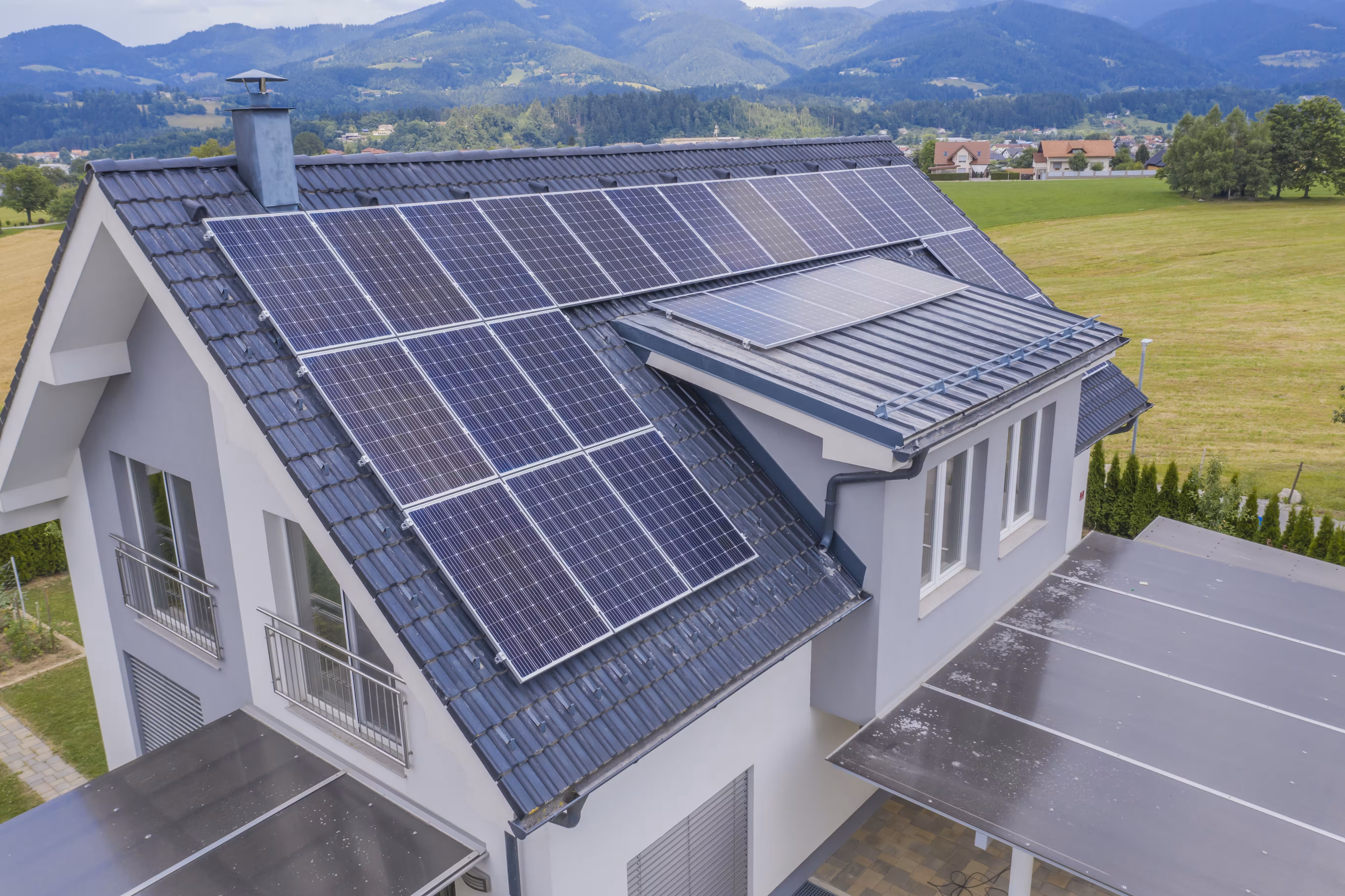 A white house with multiple solar panels on its dark gray tiled roof under a cloudy sky.