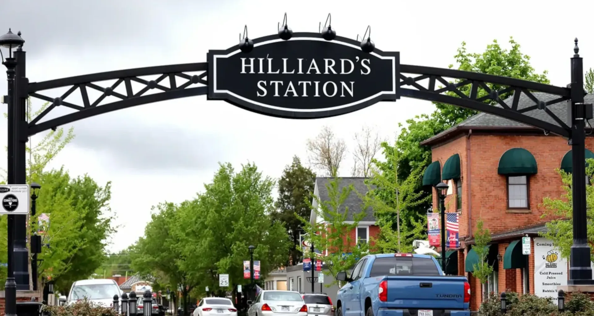 Street view of Hilliard's Station sign arch over a road with cars and brick buildings with green awnings.