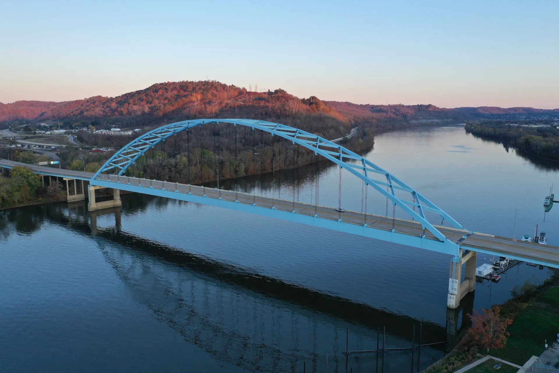 Blue bowstring arch bridge over calm river with forested hills in the background at sunset.