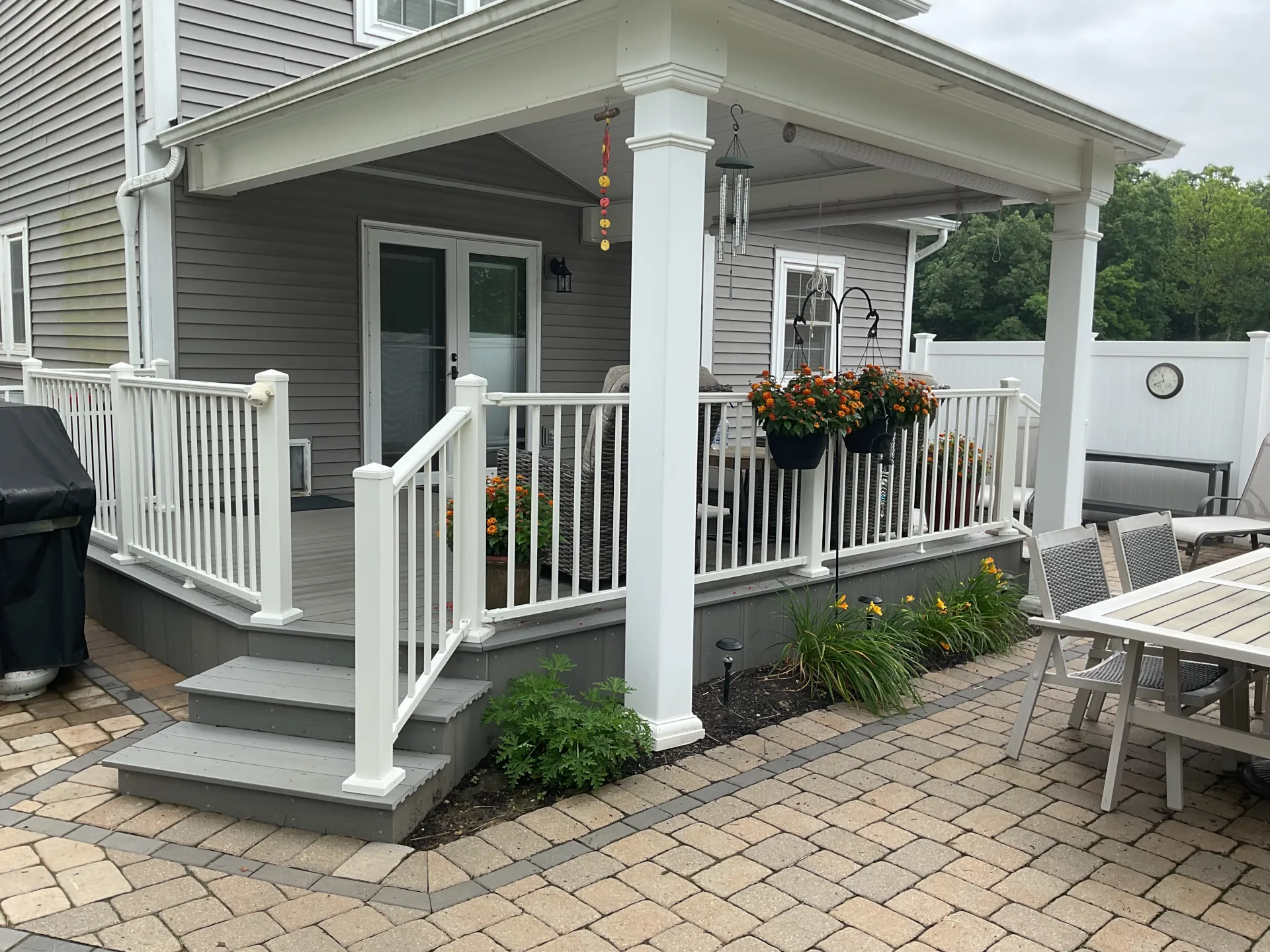 Covered back patio with white railings, steps, potted flowers, outdoor furniture, and a dining table on stone pavers.