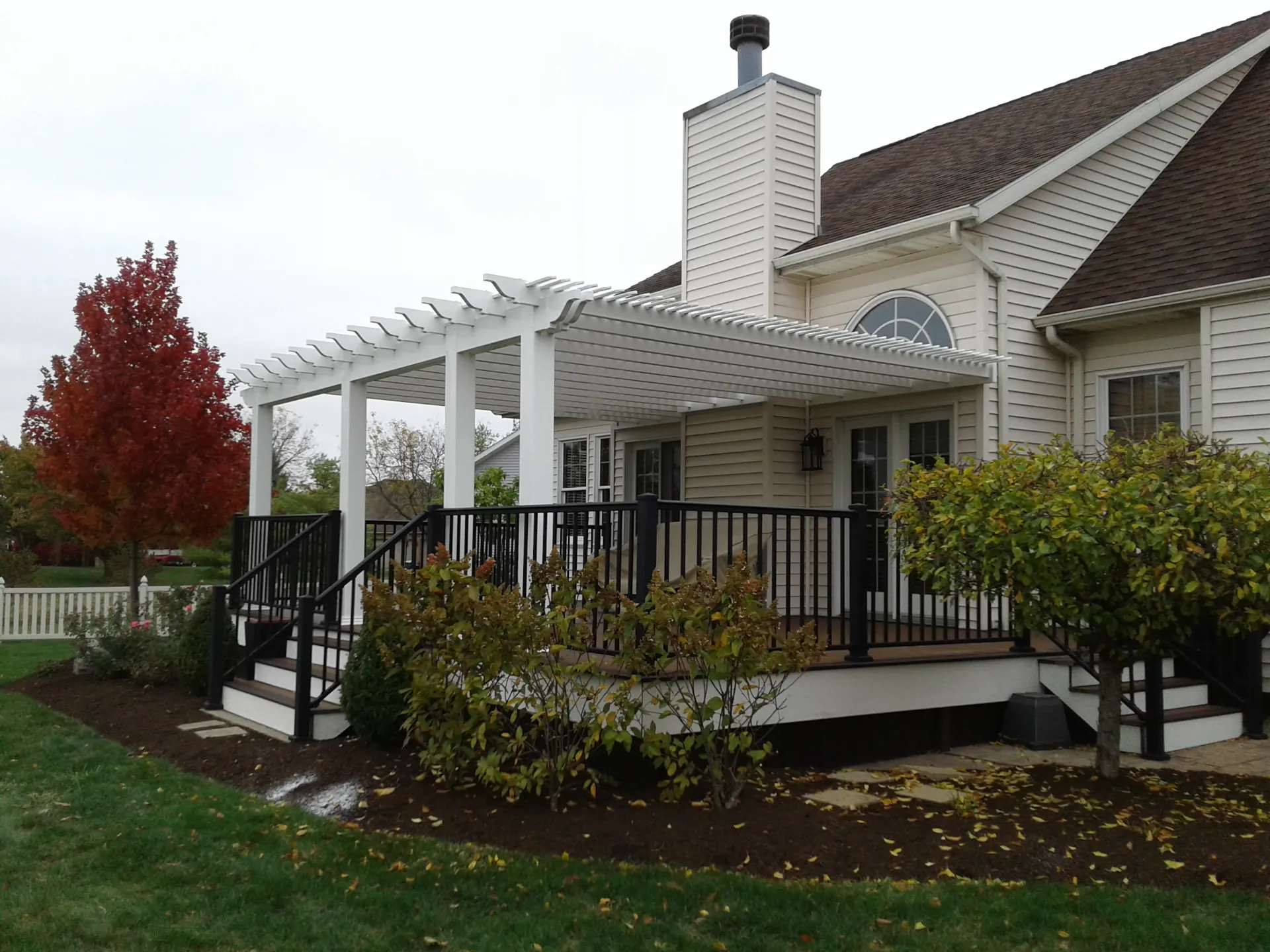 White pergola covering a back porch with black railings on a beige house with fall foliage around it.