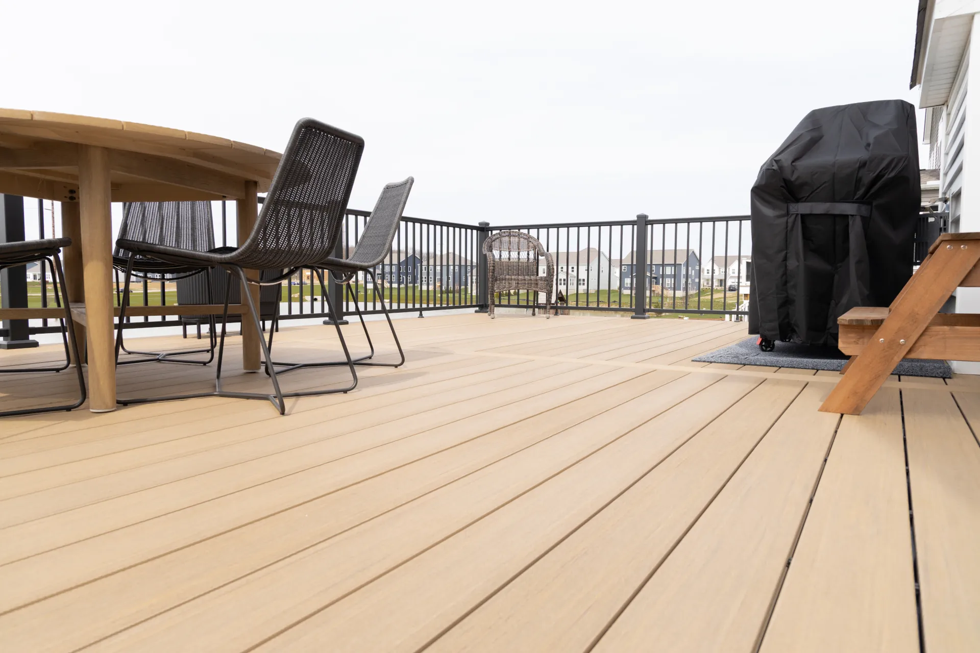 Outdoor deck with beige composite wood flooring, black metal chairs, a covered grill, and a railing overlooking homes.