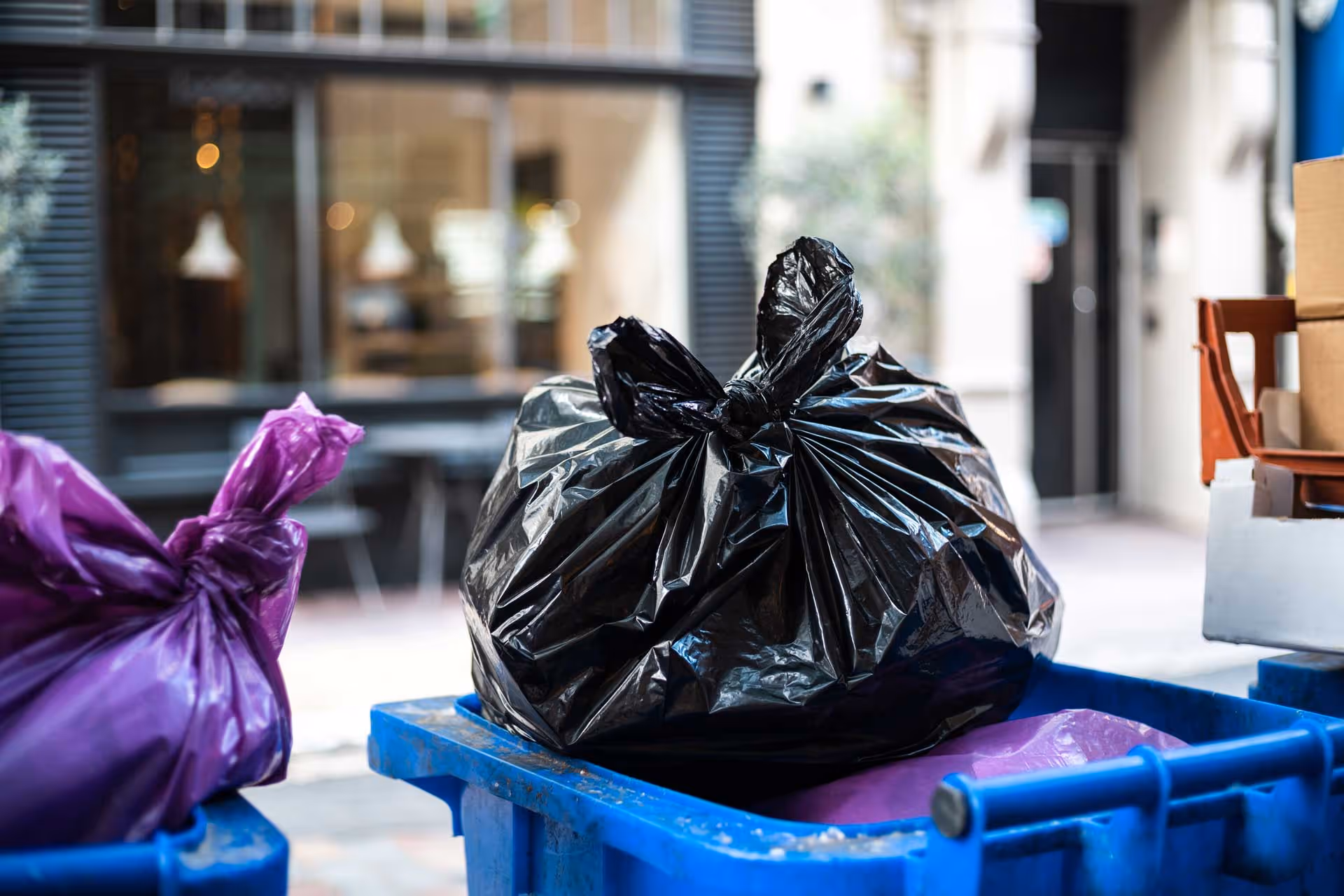 A black garbage bag sits inside a blue recycling bin on a city street.