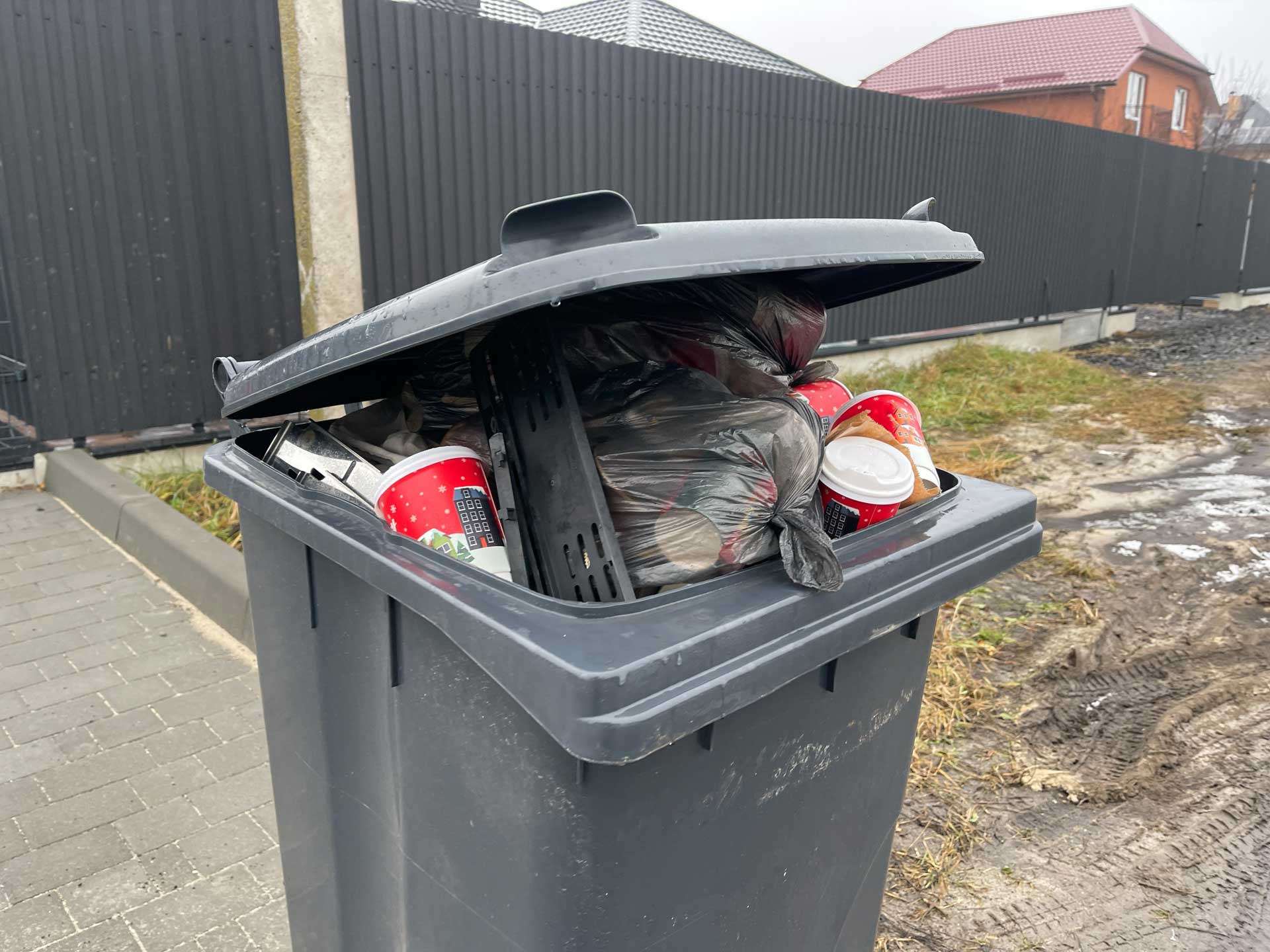 A trash bin overflowing with garbage, including paper cups and bags, stands by a fence outside.