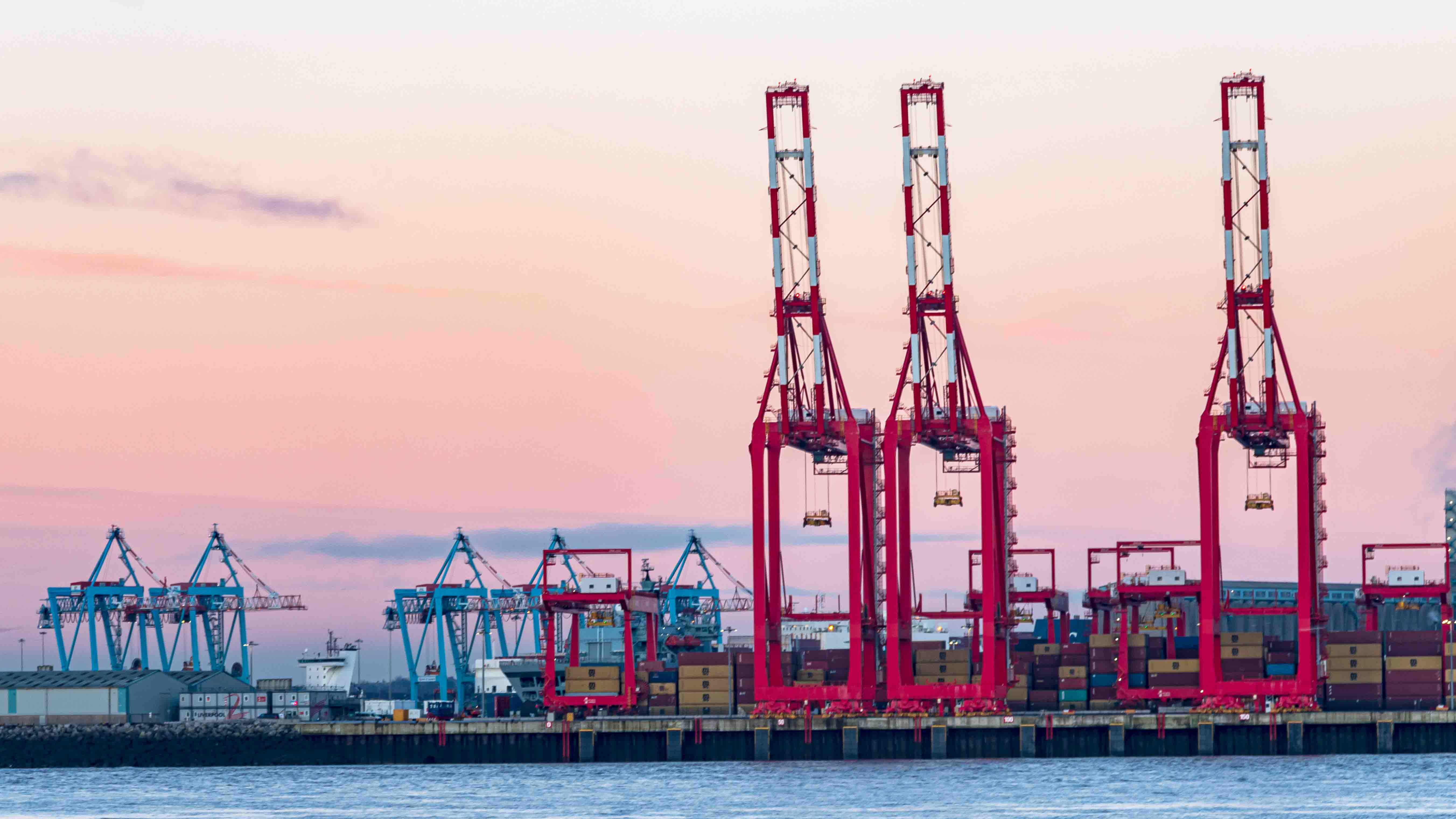 3 red shipping cranes in front of a sunset