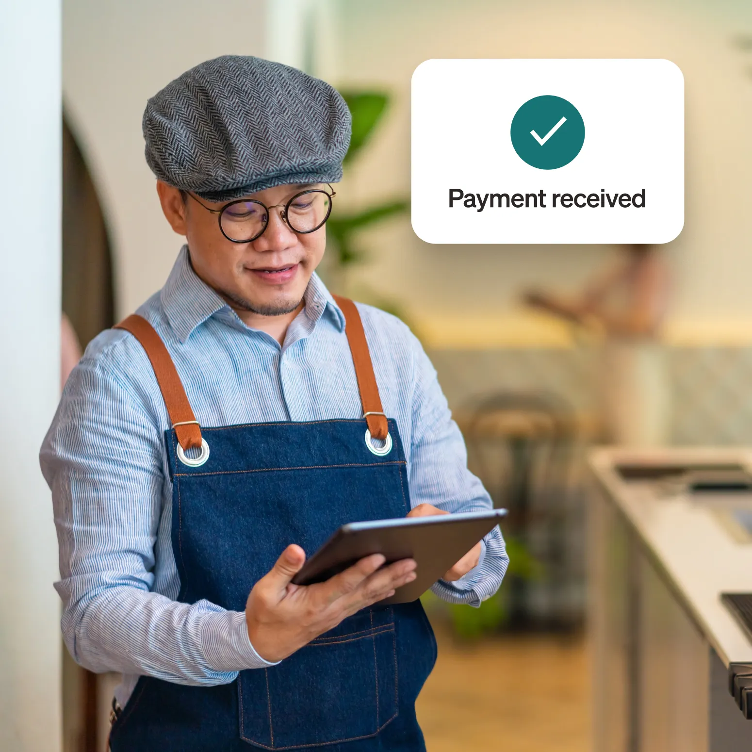 Smiling man wearing glasses and a denim apron in a cafe holding a tablet with Payment received notification.