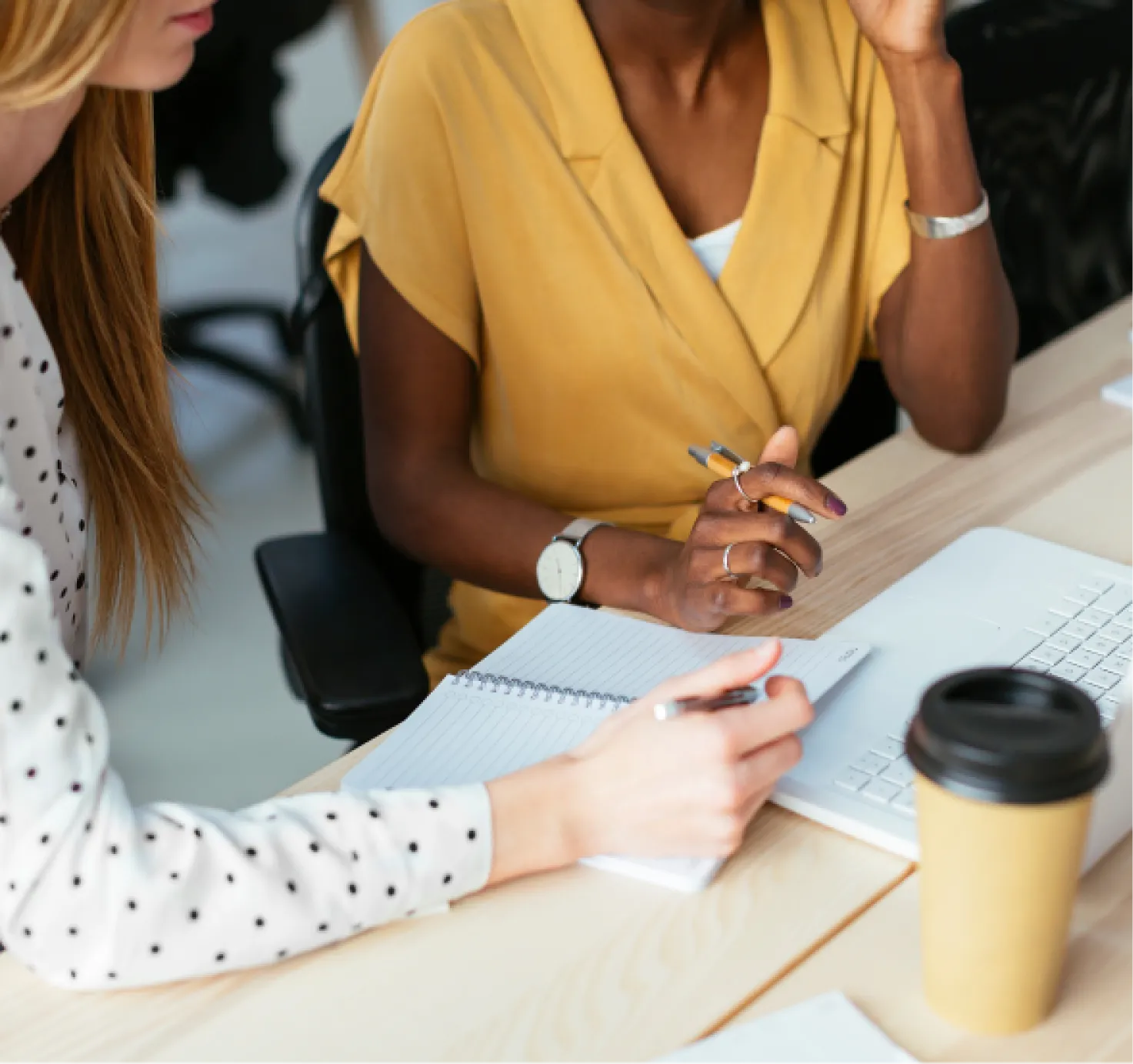 Two people are seated at a desk having a discussion over a notebook and laptop. A coffee cup sits nearby.