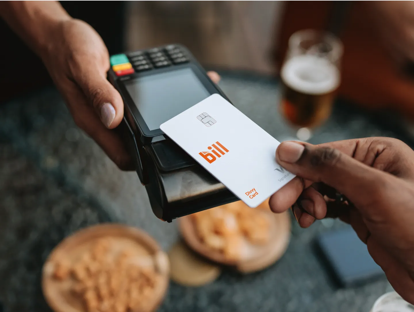 A person taps a BILL Divvy Card on a payment terminal at a table with food and a drink in the background.