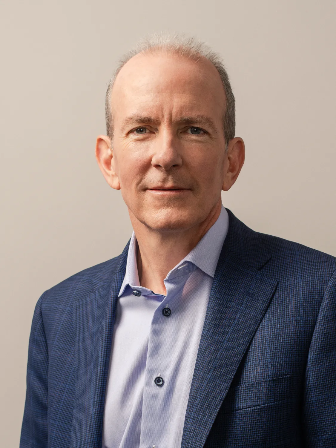 A man in a blue blazer and light blue shirt smiles against a neutral background.