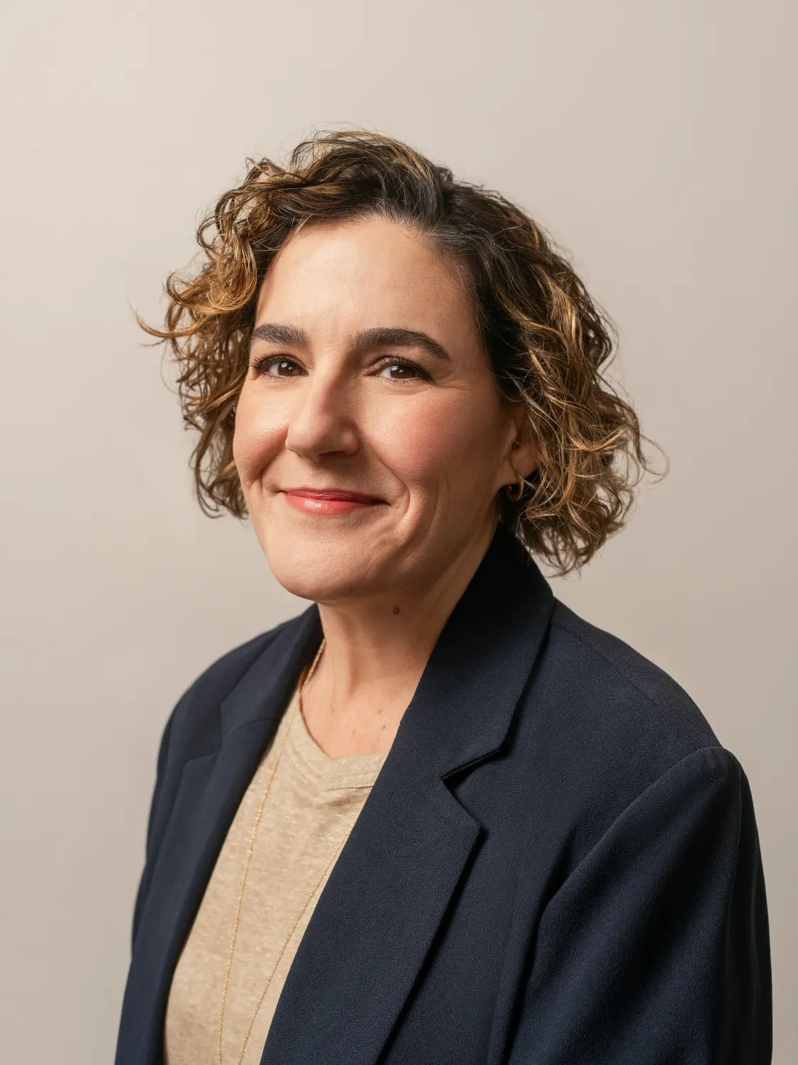 Woman with short curly hair wearing a black blazer and tan shirt smiling at the camera against a neutral background.